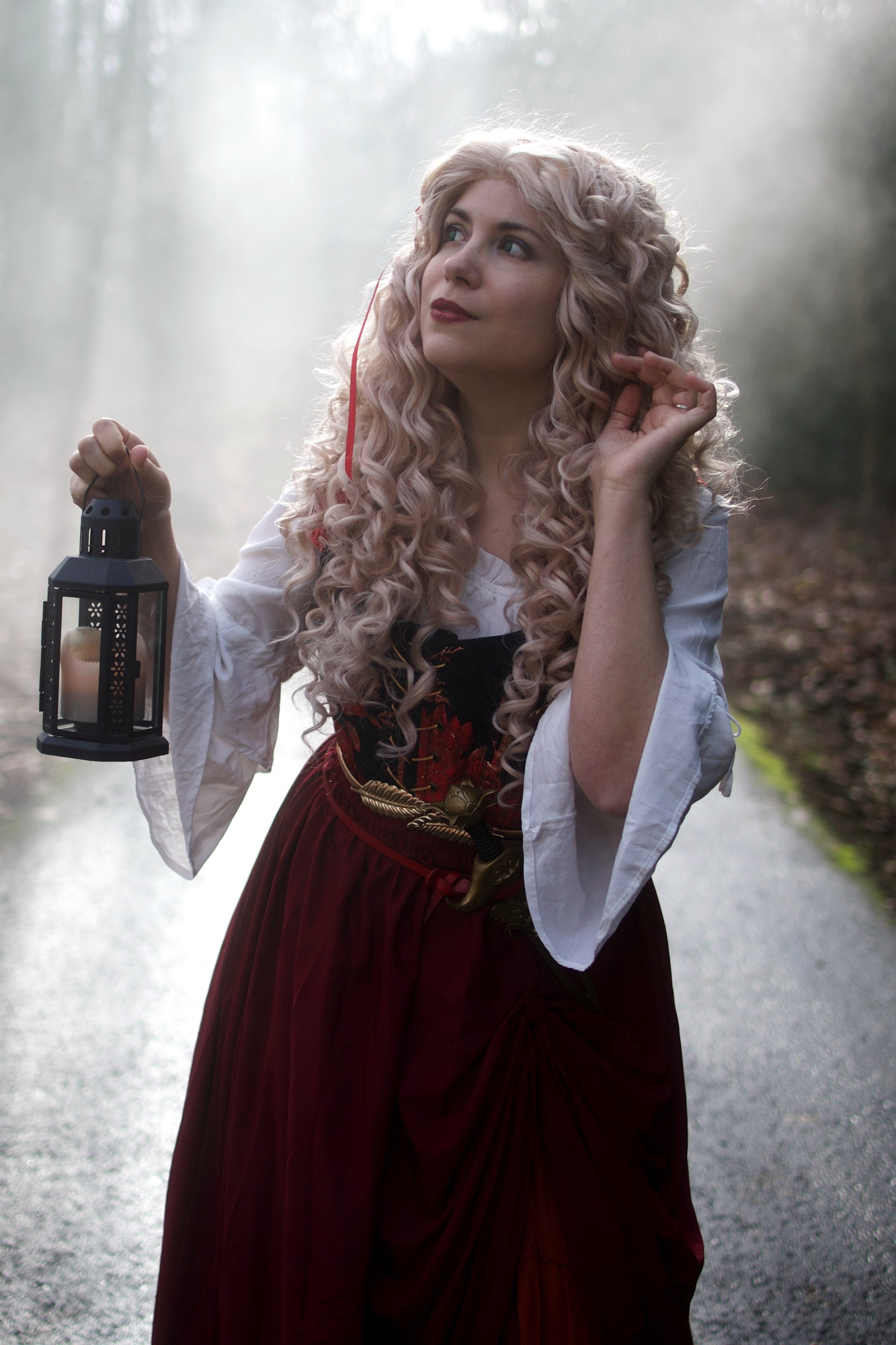 A woman in cottagecore red skirt, black and red floral corset, and white blouse. She is holding up a candle in a lantern and staring off to one side, surrounded by mist.