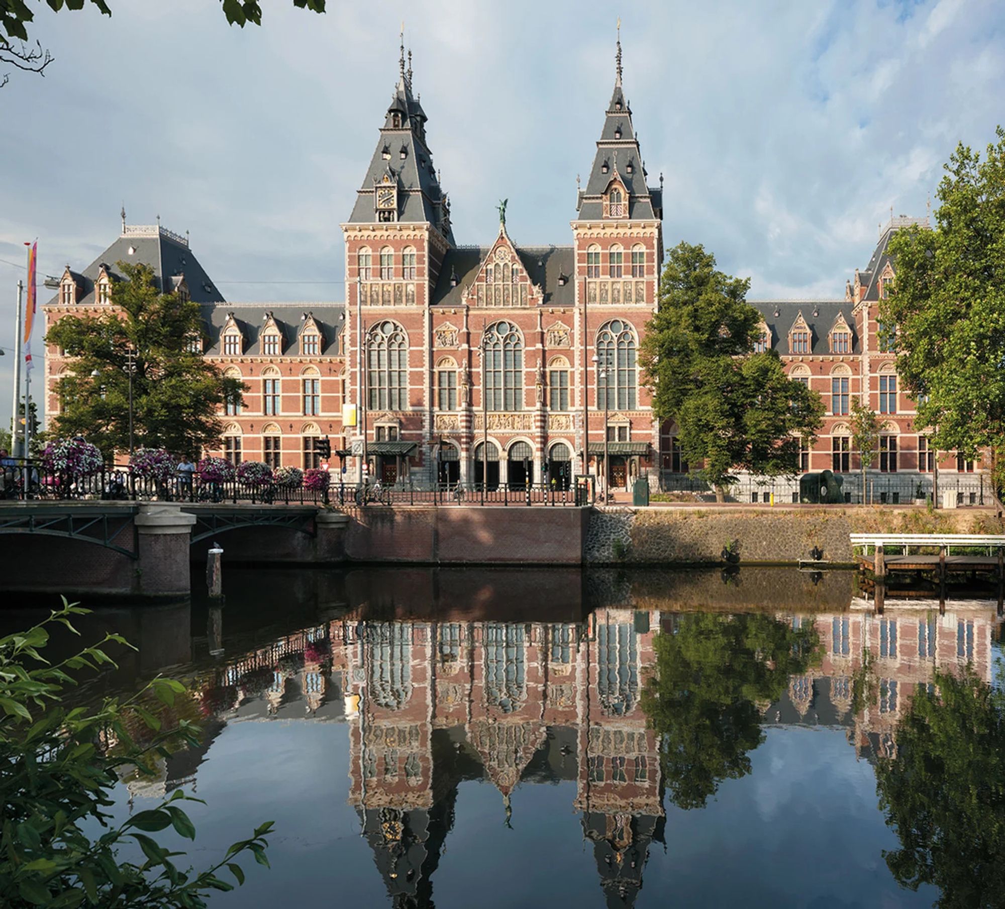 Image shows the front of the Rijksmuseum in Amsterdam, a gothic-style, red-brick facade; the facade is reflected in the water of the canal in the foreground.