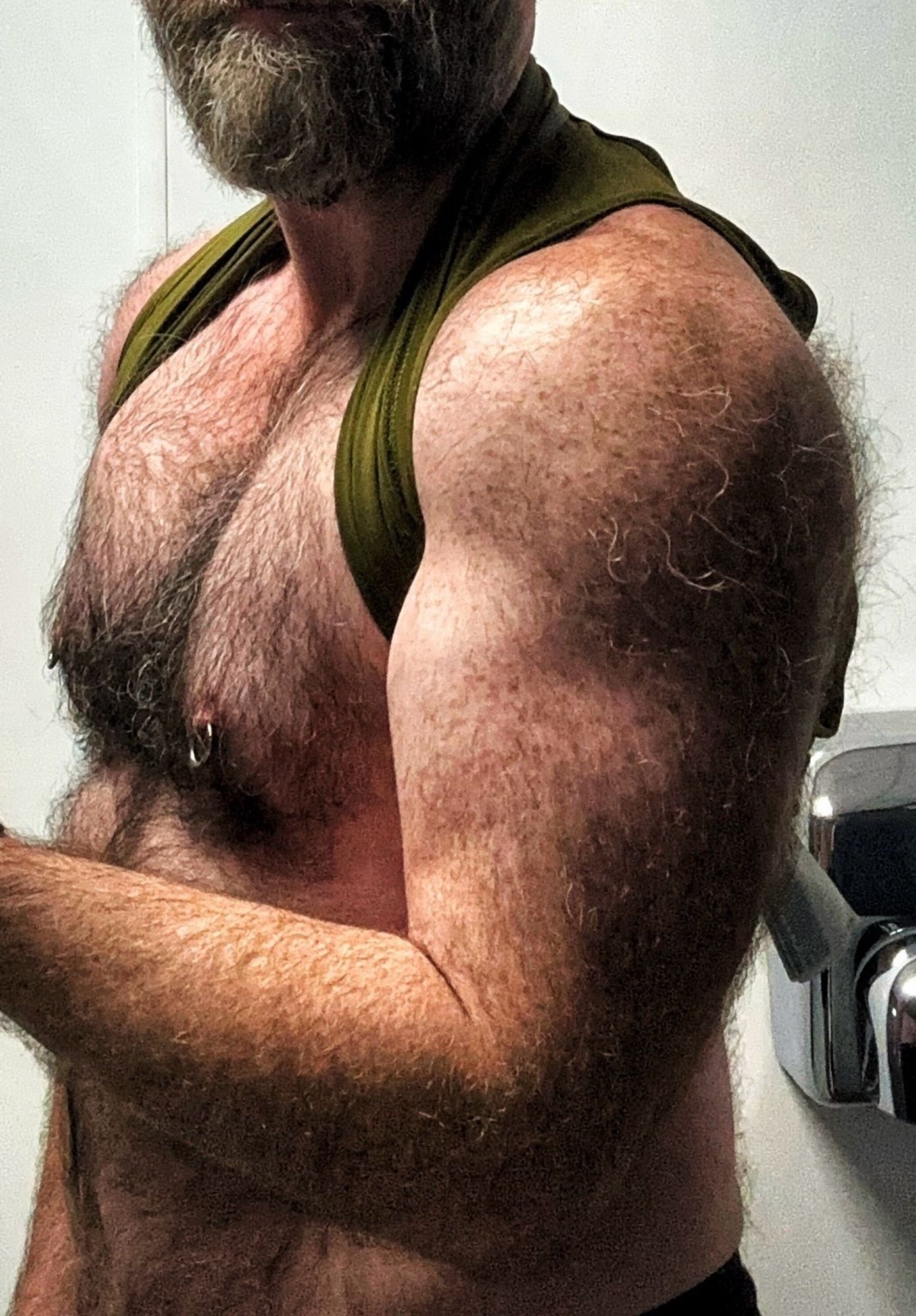 A hairy arm flexing after a gym workout. Vest is lifted to show hairy chest and pierced nipples.