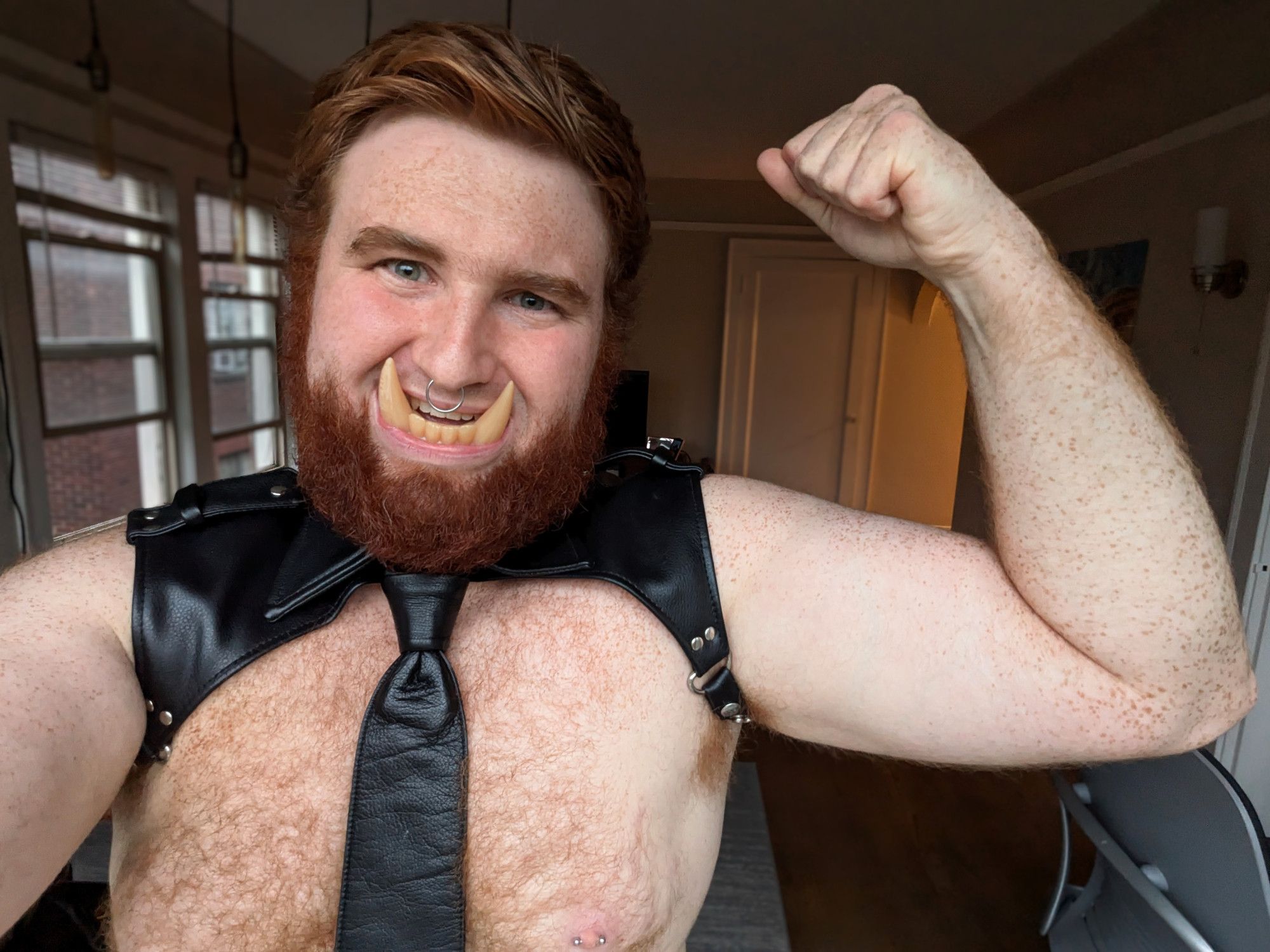 A bearded red-haired man in a leather harness and tie with molded orc tusks in his mouth. He is standing and flexing his bicep, and smiling while baring his teeth at the camera. #pinup