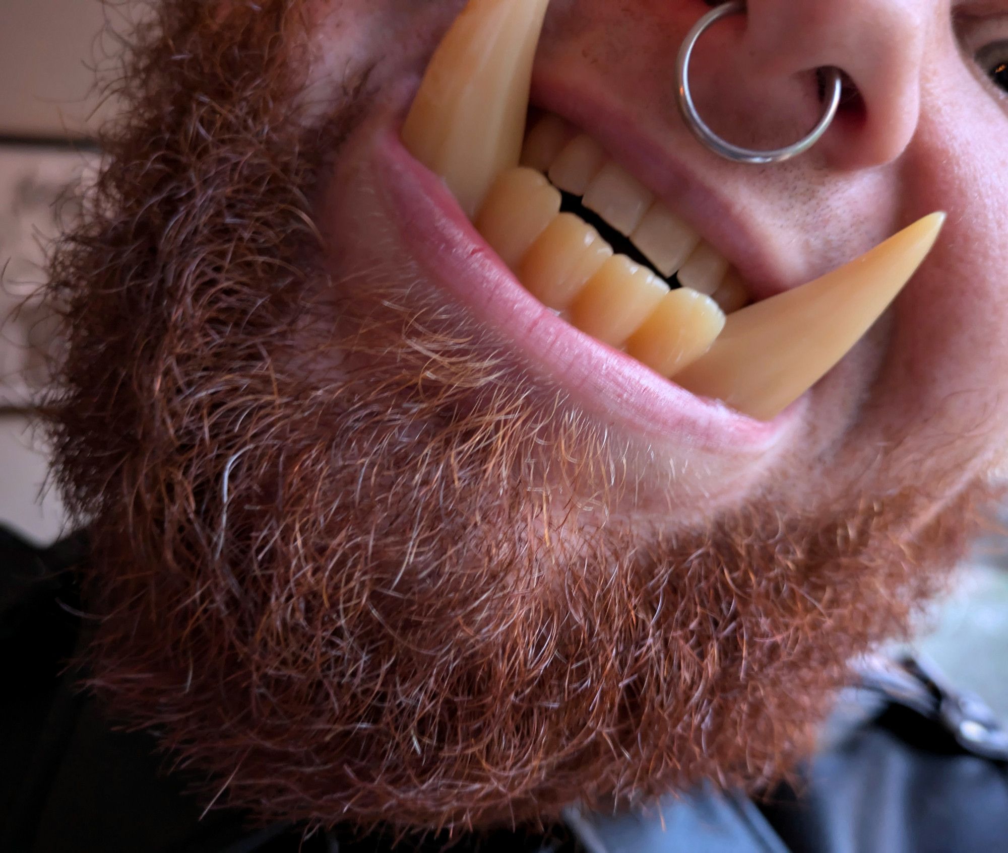 An extreme close-up of a man with a red beard and molded orc tusks in his mouth. The camera is focused on his teeth, lips, and beard.