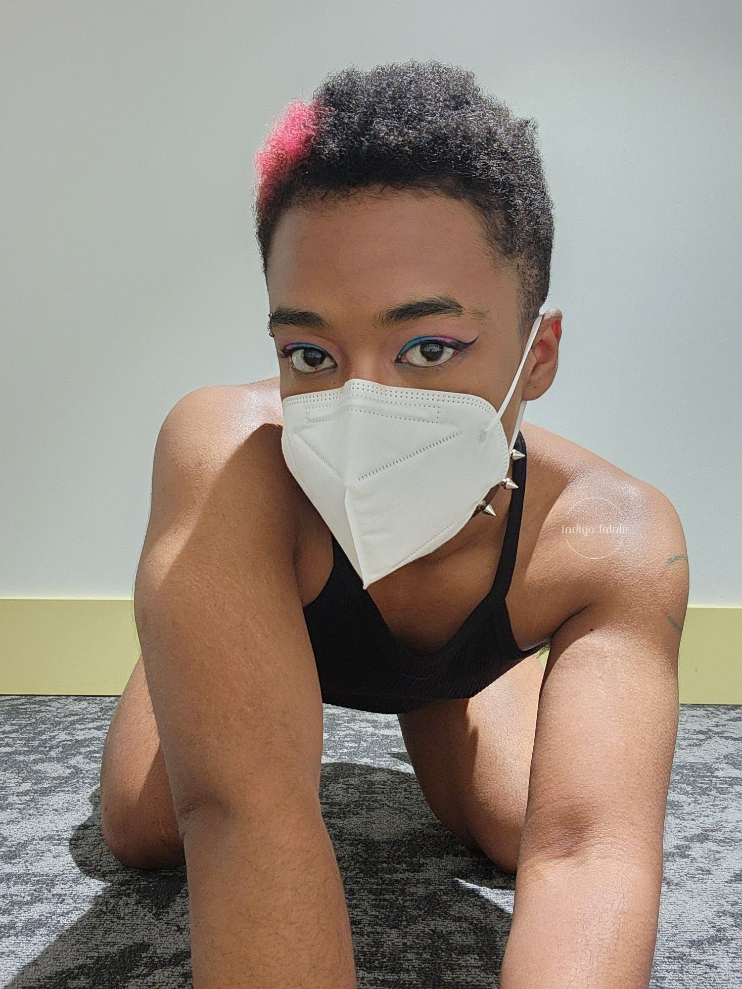 Indigo is kneeling on all fours. It's wearing no glasses, a white KN95 mask, a black tank crop top and black jean shorts with eyeshadow that matches the bisexual flag.