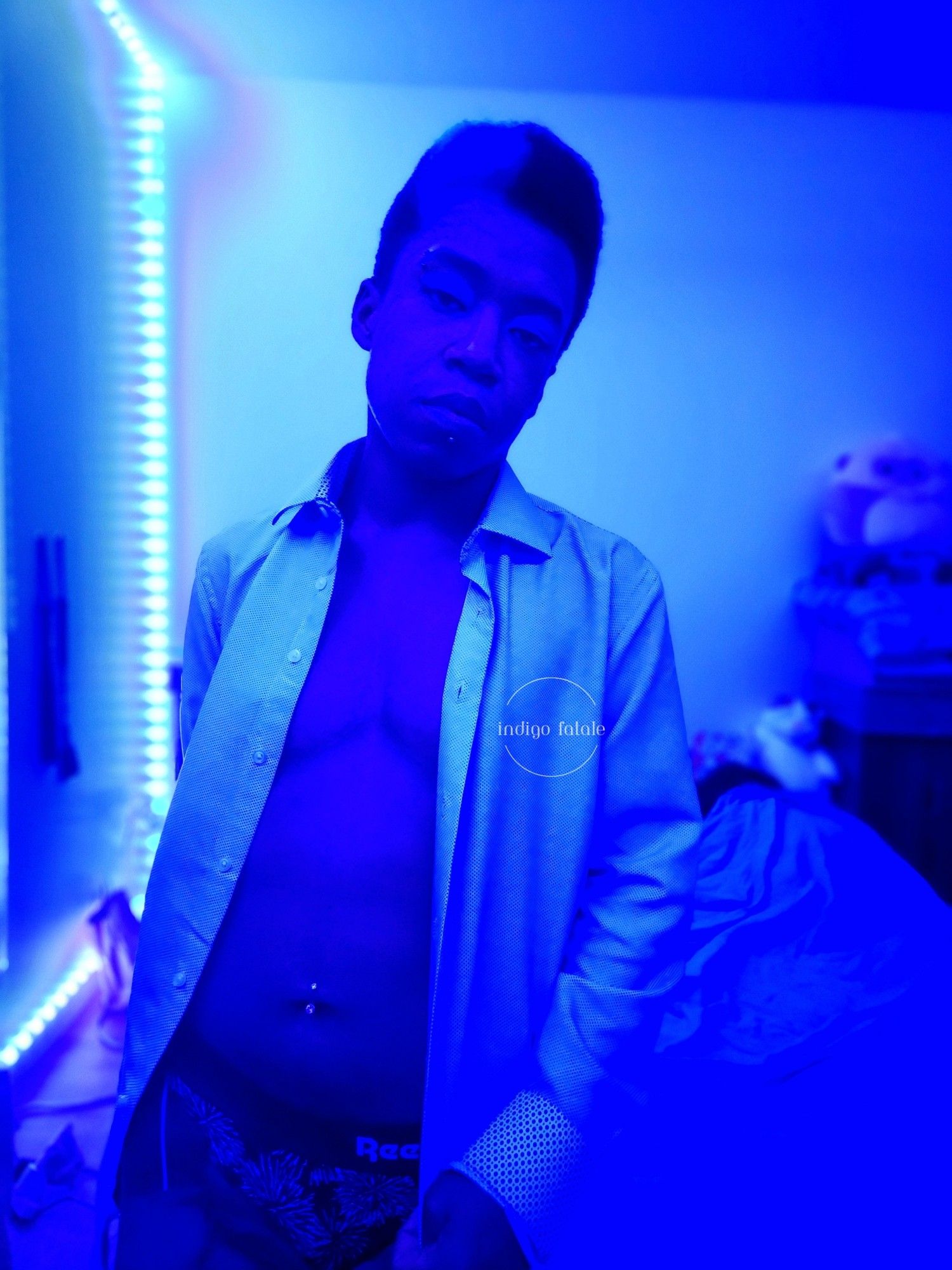 Indigo in a white collared shirt that's open and showing its chest, a black and white thong and blue eyeshadow with blue lighting.