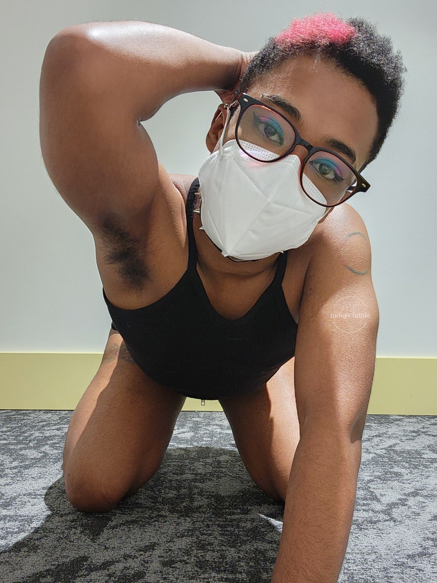 Indigo is kneeling with one arm up and the other on the floor. It's wearing its glasses, a white KN95 mask, a black tank crop top and black jean shorts with eyeshadow that matches the bisexual flag.