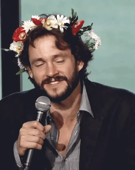 Hugh Dancy speaking into a microphone and laughing while wearing a red-and-white flower crown