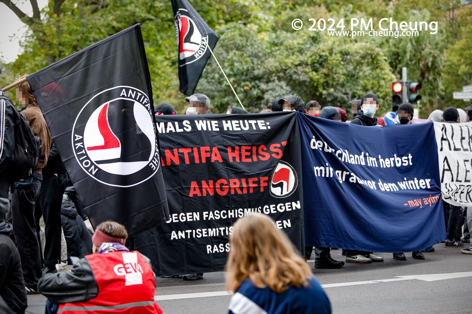 Bei der Zwischenkundgebung am Sportkomplex an der Rennbahnstraße sind mehrere Banner und Fahnen zu sehen. Auf einem der Banner ist "Damals wie heute: Antifa heißt Angriff! Gegen Faschismus, gegen Antisemitismus, gegen Rassismus!" zu lesen.