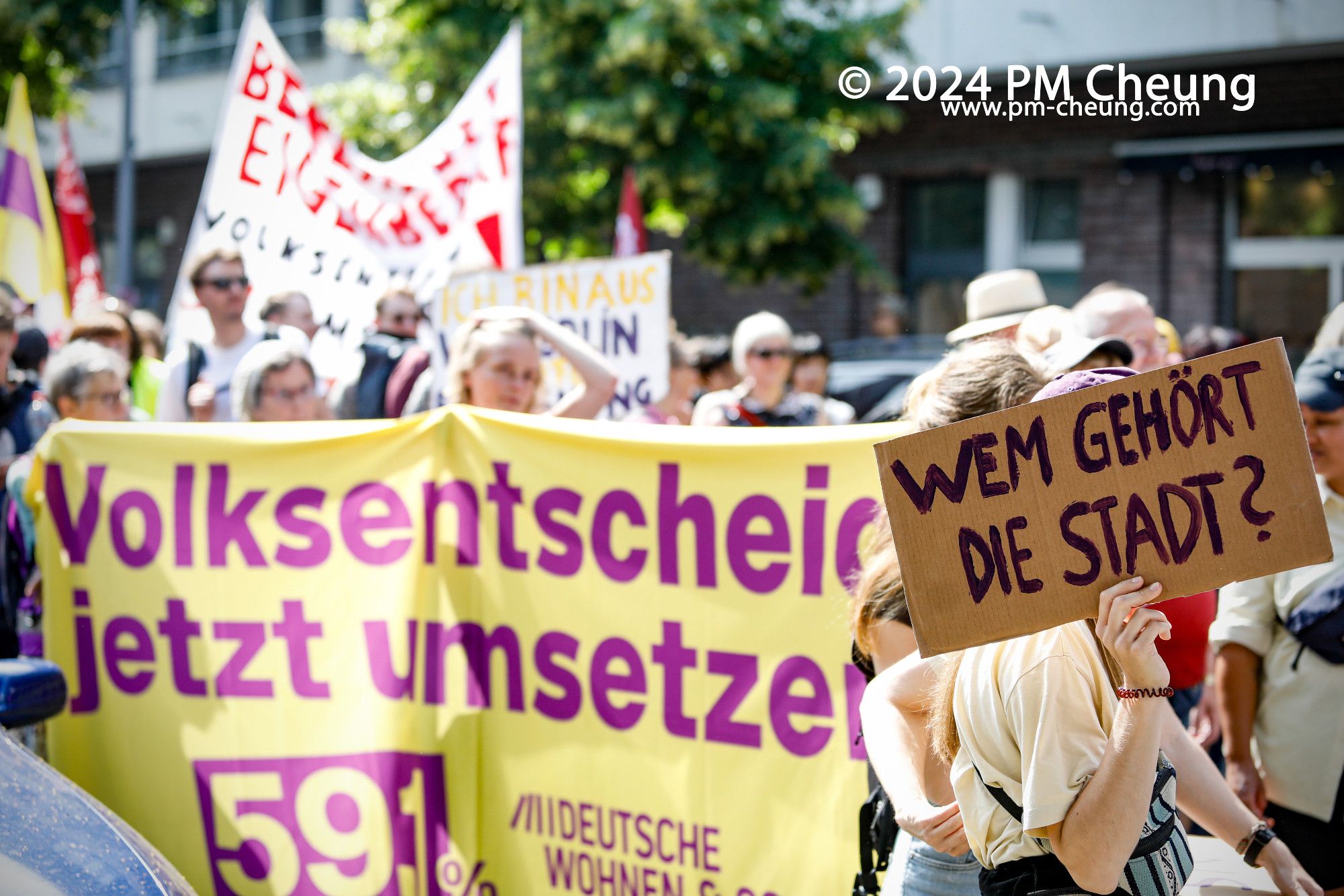 Auf der Schöneberger Straße stoppte der Aufzug nahe dem Anhalter Bahnhof für eine Zwischenkundgebung. Hier ist unter anderem eine Demonstrant*in mit einem "Wem gehört die Stadt" Schild zu sehen. Im Hintergrund ist ein Banner der Initiative "Deutsche Wohnen & Co" zu sehen.