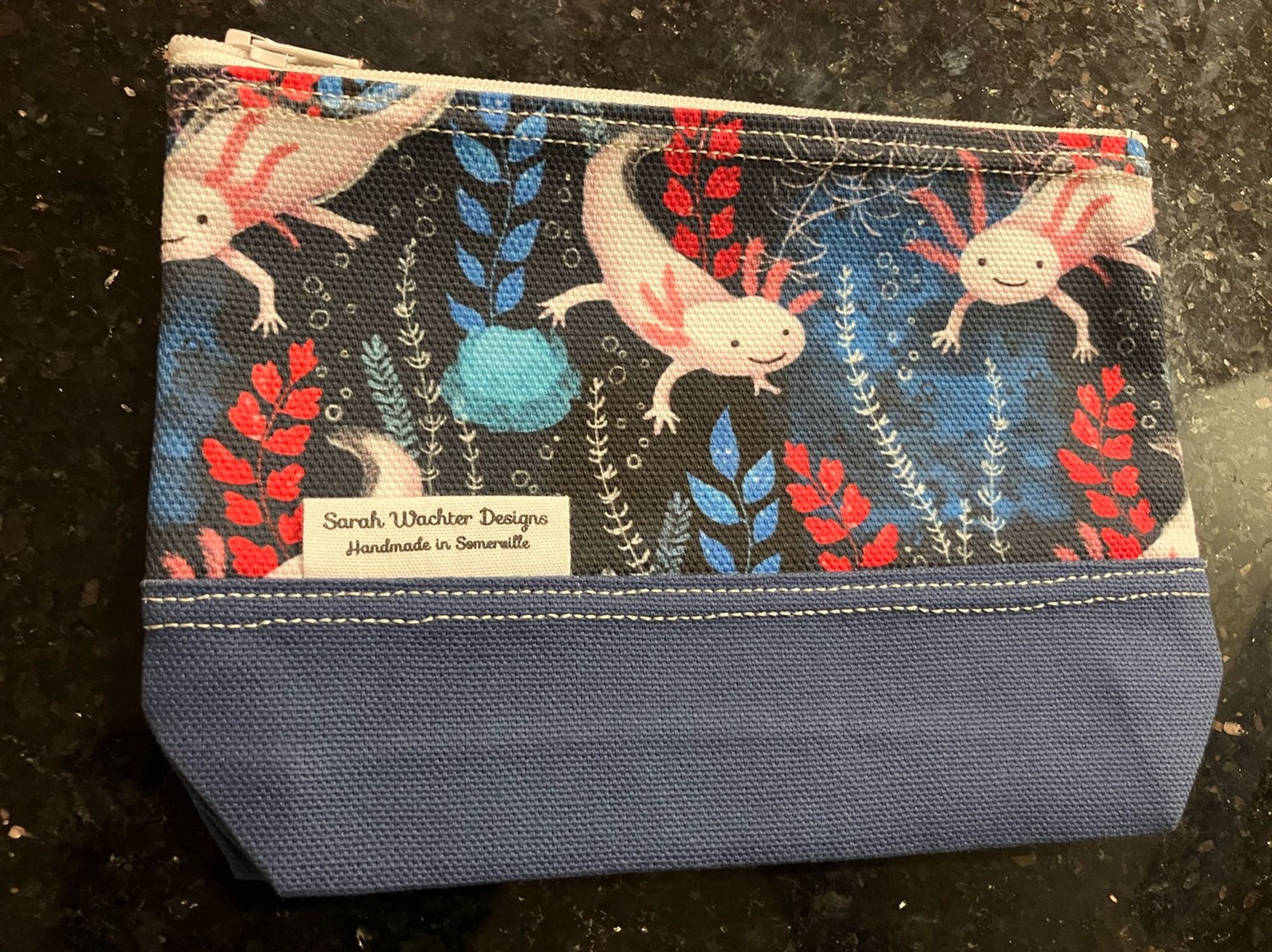 A small zippered pouch with an axolotl-print fabric. There’s a tag on the pouch resting “Sarah Wachter Designs, handmade in Somerville”