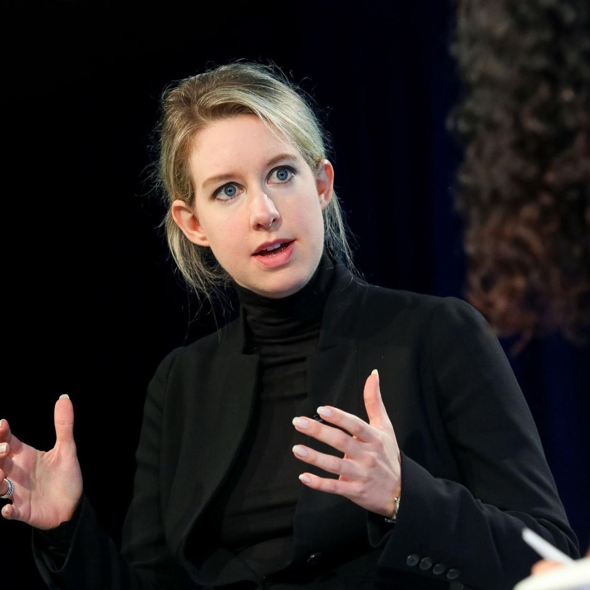 A photo of Elizabeth Holmes wearing a black turtleneck and black blazer. It seems to be taken in an interview before her fraud was revealed since she's channeling Steve Jobs vibes.