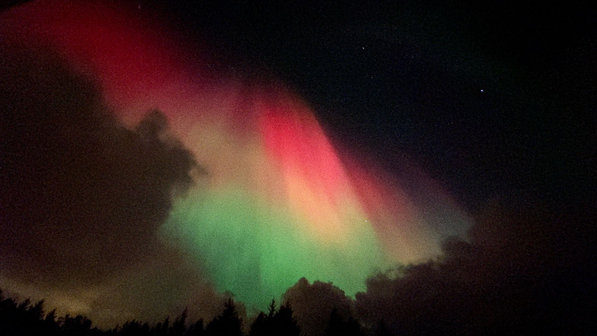 Aurora that’s red on top and green at the bottom and orangey in between. The red goes from the top left corner of the photo and down across towards the bottom right corner. It’s partially obscured by clouds.