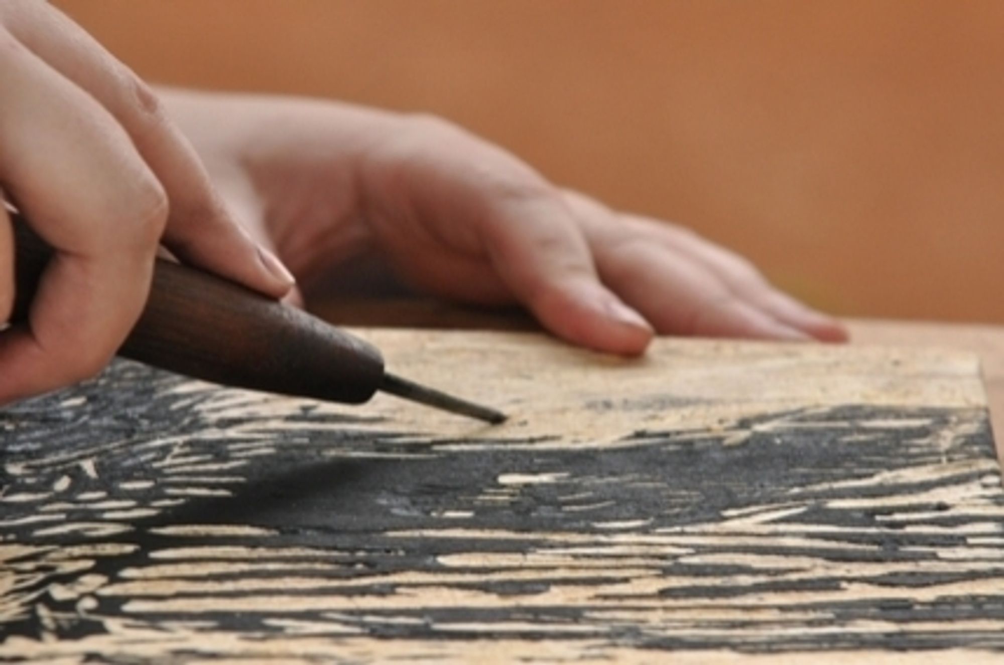 Mão direita segura uma ferramenta de xilogravura,  goiva, contra uma placa de madeira com manchas de tinta preta, a Mão esquerda segura a placa