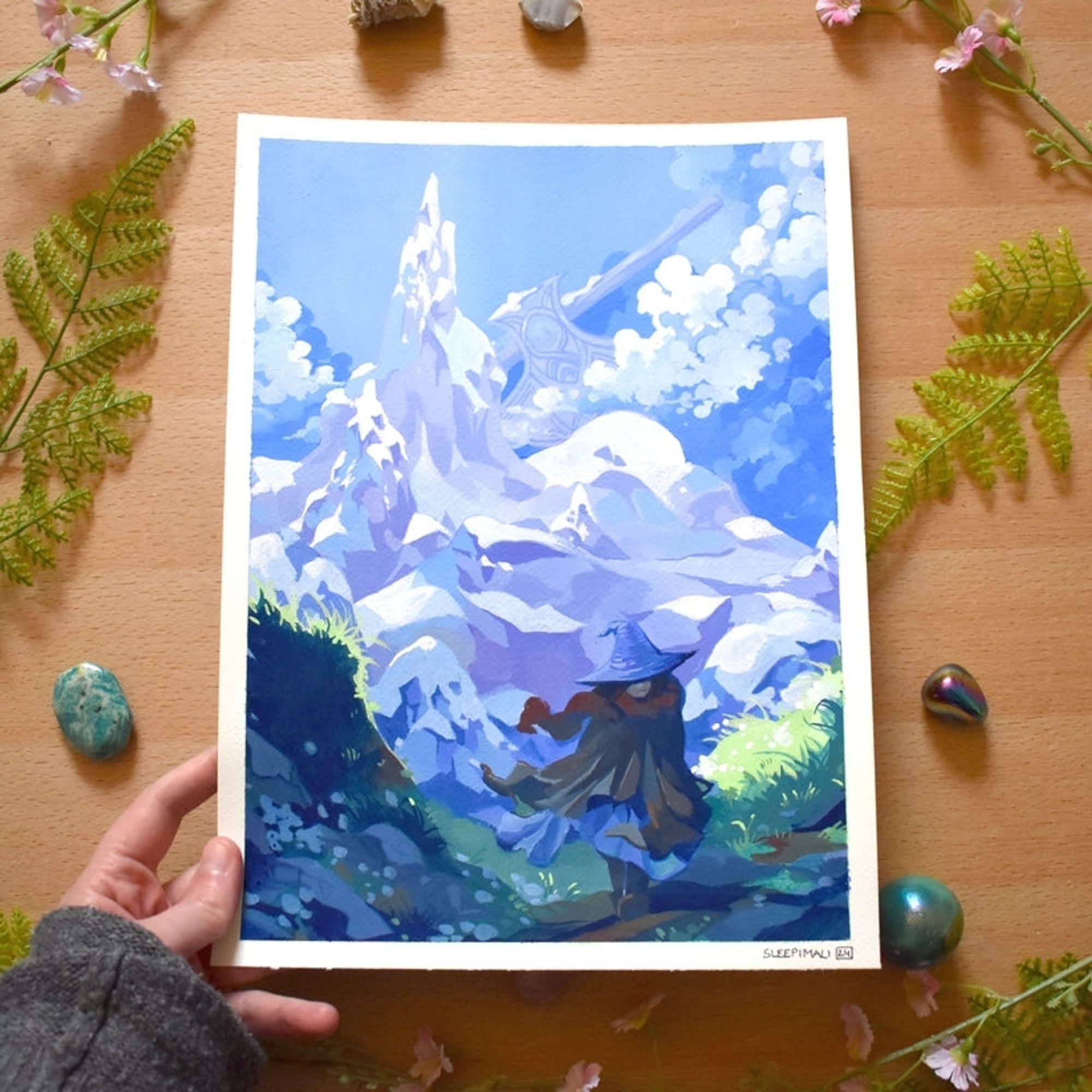 Photo of my hand holding a painting of a person in a witch hat, red scarf and warm clothes blowing in the wind. In the background there's a tall, sharp mountain with a giant axe stuck in its side