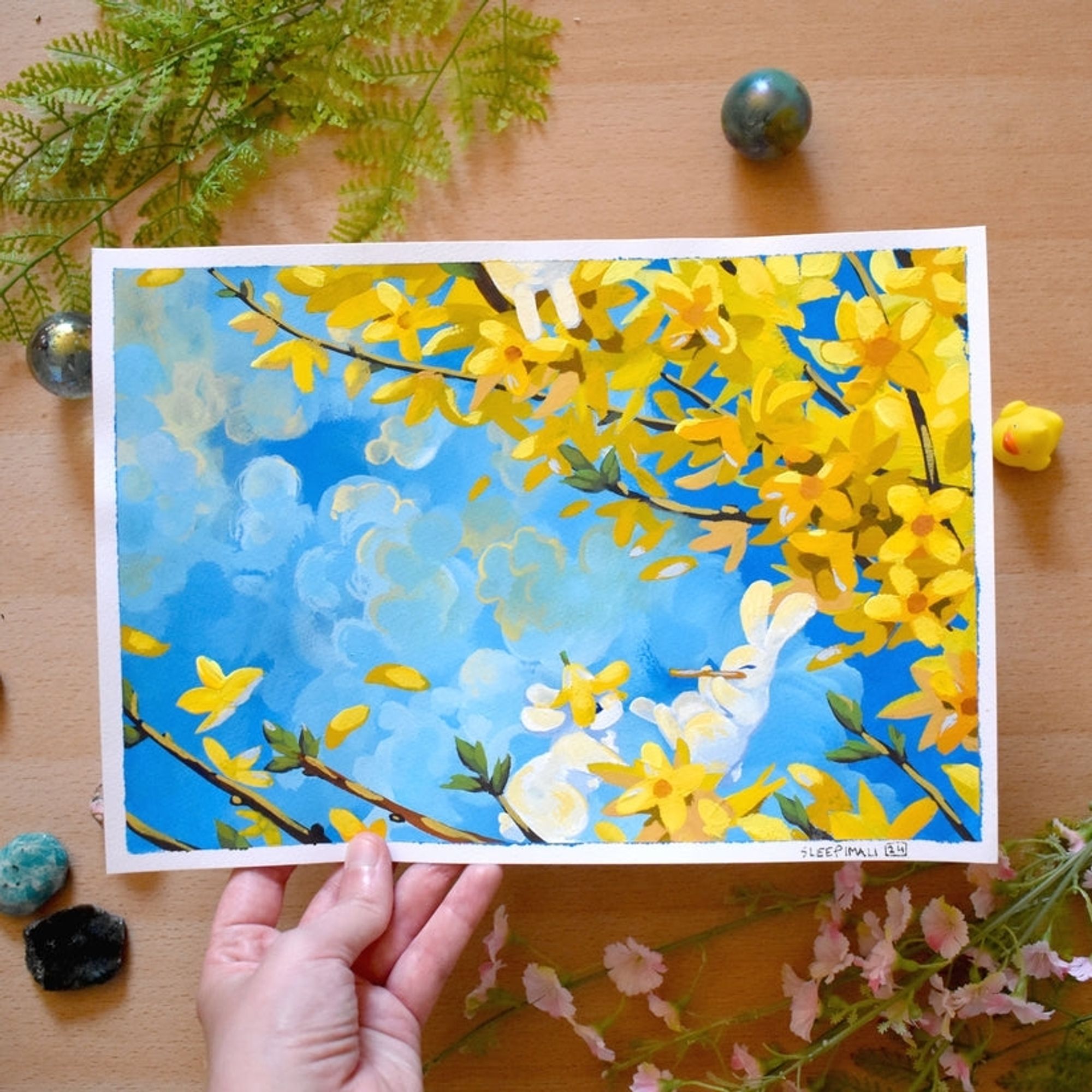 Photo of an original acrylic gouache painting of white bunnies on bright yellow forsythia flowers against a blue sky with soft clouds. One of the bunnies is wearing a forsythia flower hat and listening to another bunny happily playing the flute. A couple of bunny legs also dangle from the top of the painting