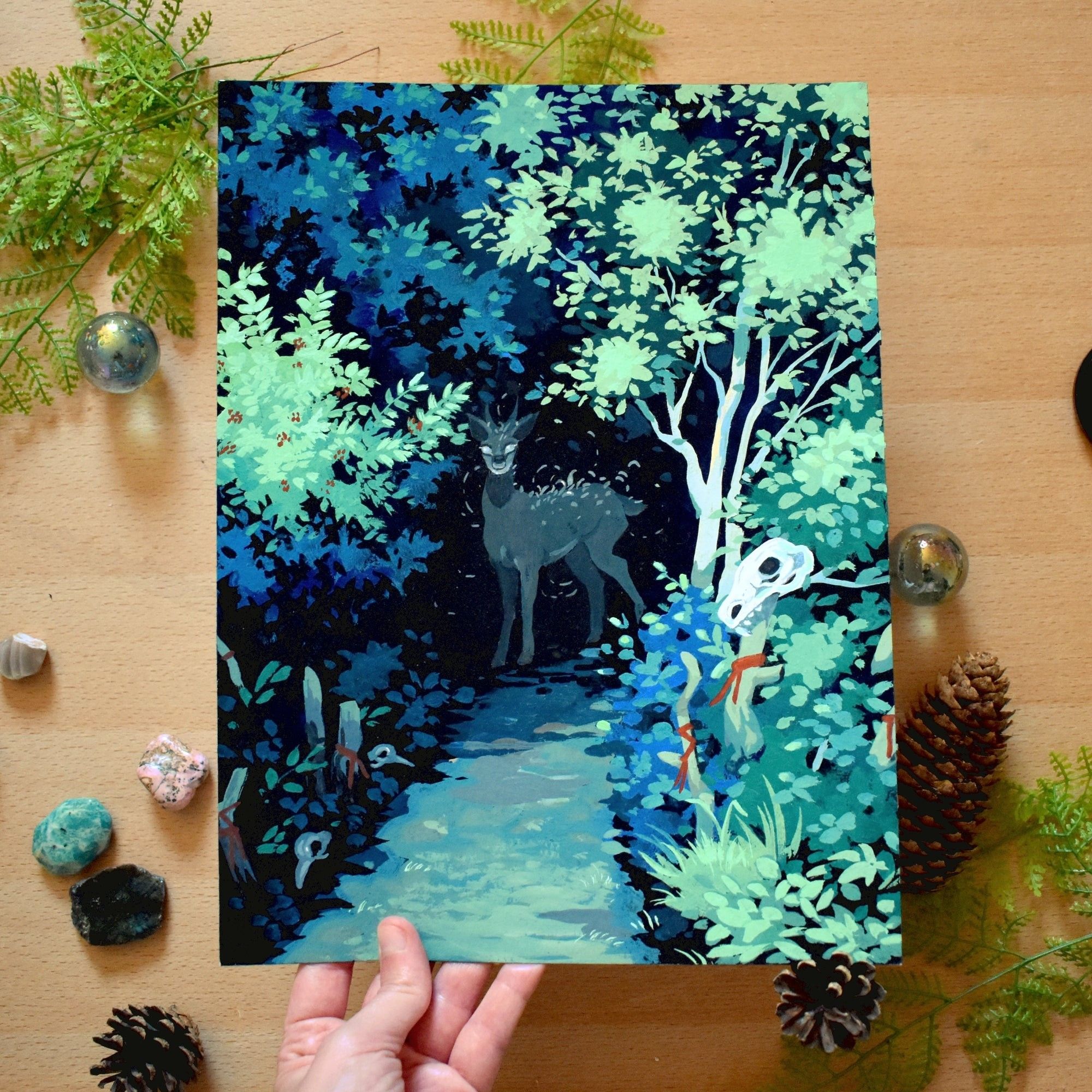 Photo of an acrylic gouache painting of a forest path leading into a tunnel of trees where the light doesn't reach. At the opening of the tunnel, there is a deer-like figures with glowing bright eyes and some type of vegetation growing on its back. In the area leading  up to the tunnel there are a few sticks tied with red ribbons and on one of them there's a skull of an unknown animal. There are also a couple of bird skulls on the ground.