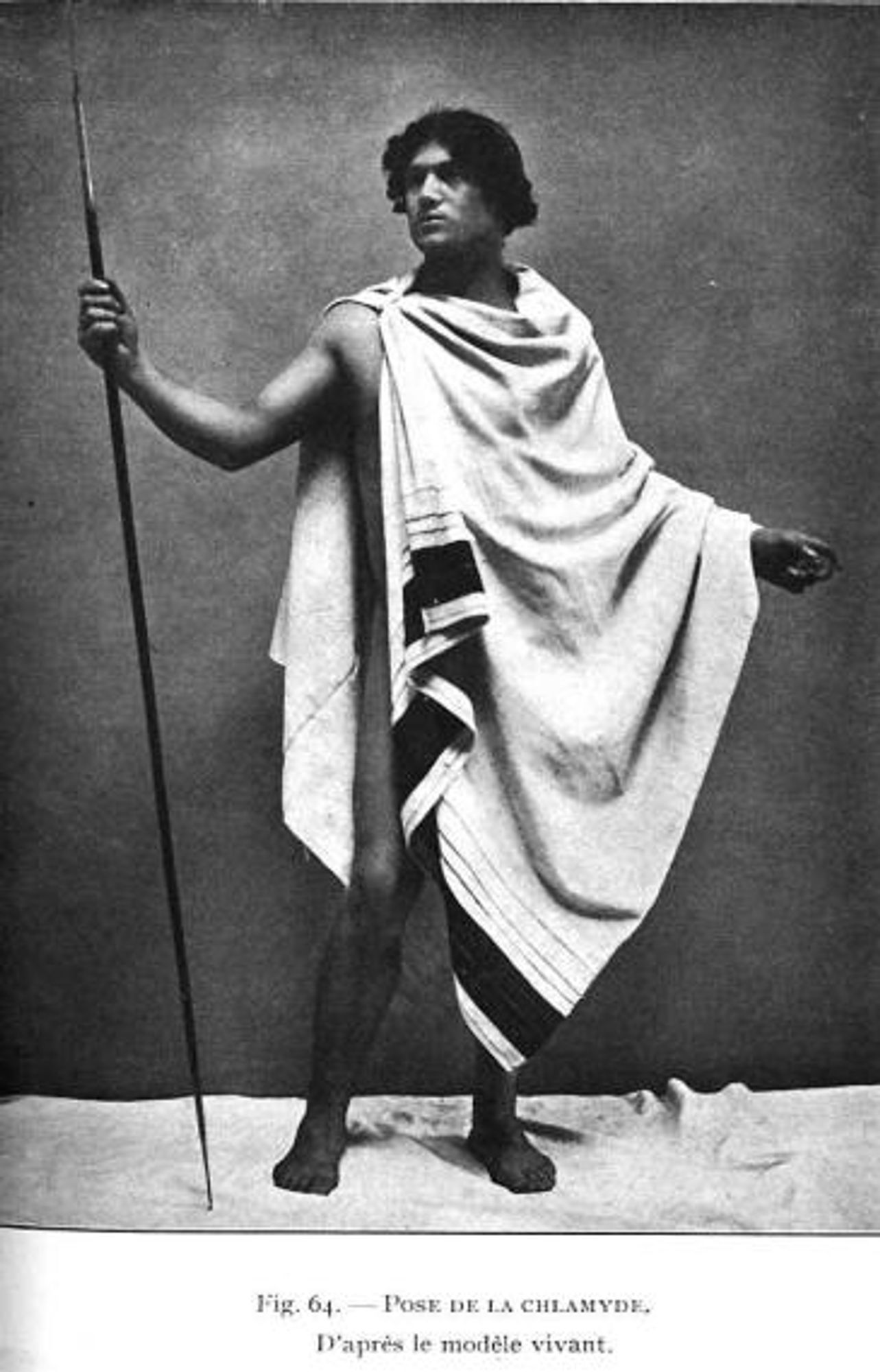 old black and white photo of a model wearing a chlamys, a type of ancient greek cloak, and holding a spear