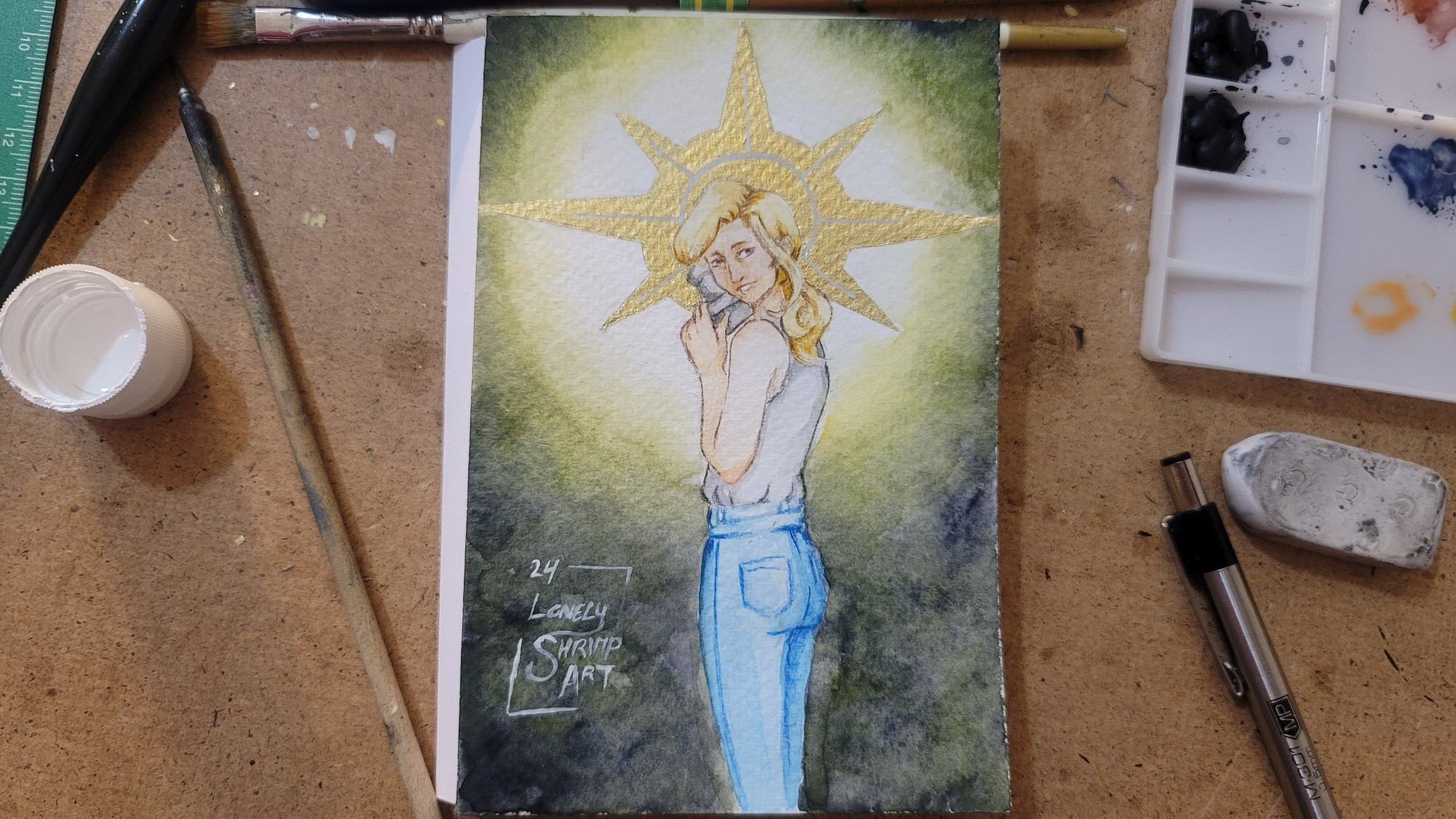 A watercolor painting of Alice Wake, from the Alan Wake series. She is a white woman who stands facing away from the viewer, in blue jeans and a grey tank top. Her blonde hair is tied back in a ponytail, and she has a smile on her face as she looks back towards the viewer. she has a camera in her hands. 
Behind her head is a golden sun-like halo, seemingly cutting through the grey-green darkness surrounding her.
The painting sits on a brown table, with a various art supplies surrounding it.