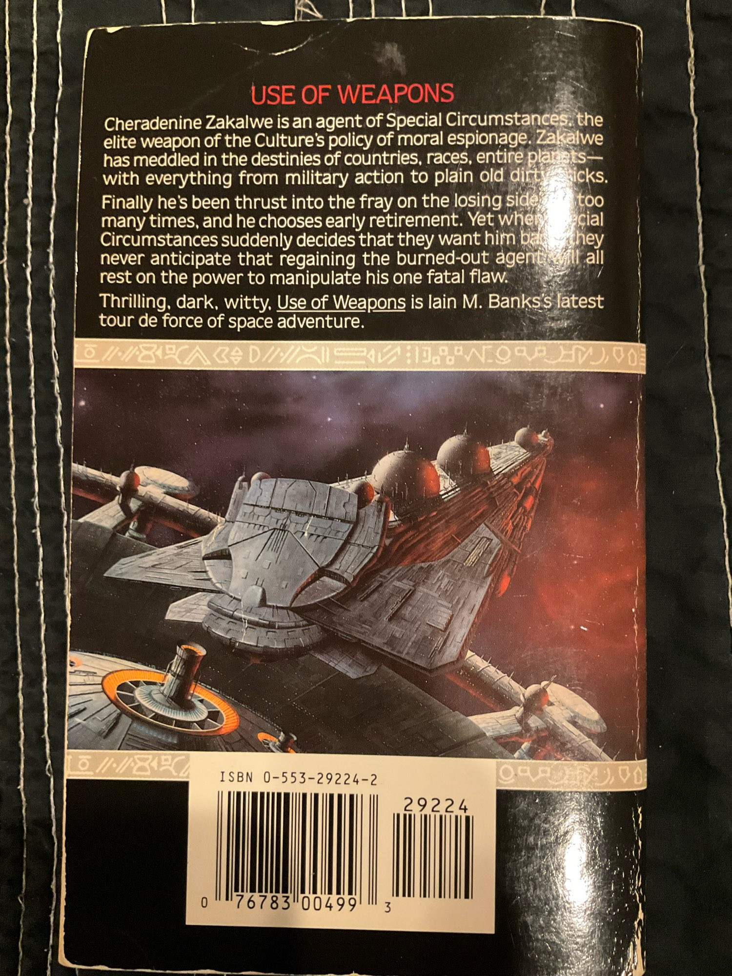 the back cover of a well-loved mass market paperback edition of Iain M. Banks' Use of Weapons.

USE OF WEAPONS

Cheradenine Zakalwe is an agent of Special Circumstances, the elite weapon of the Culture's policy of moral espionage. Zakalwe has meddled in the destinies of countries, races, entire planets -- with everything from military action to plain old dirty tricks.

Finally he's been thrust into the fray on the losing side too many times, and he chooses early retirement. Yet when Special Circumstances suddenly decides that they want him back, they never anticipate that regaining the burned-out agent will all rest on the power to manipulate his one fatal flaw.

Thrilling, dark, witty, Use of Weapons is lain M. Banks's latest tour de force of space adventure.

pictured: a painting of a huge spaceship, with an even more gargantuan space habitat in the background.