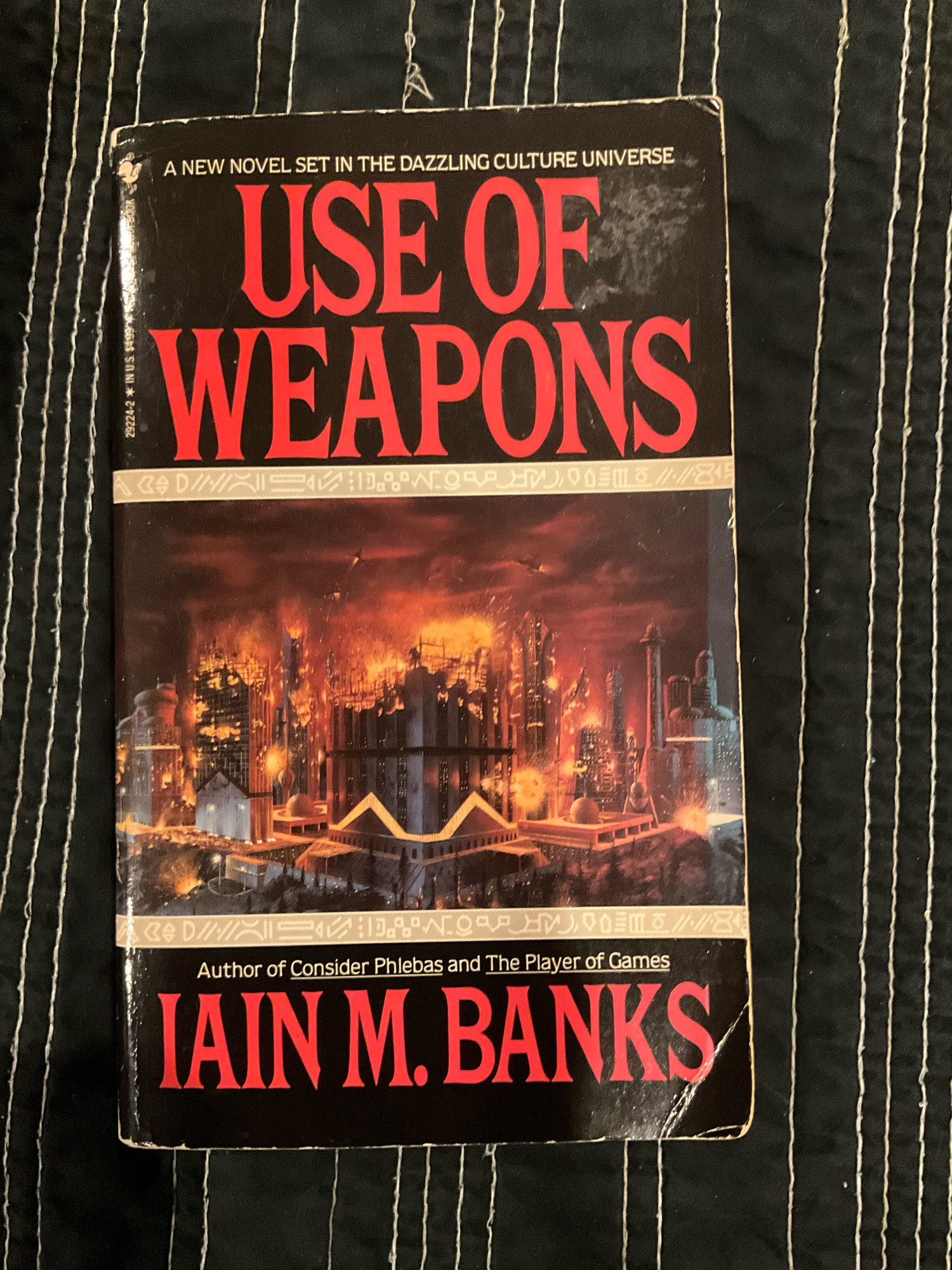 the front cover of a well-loved mass market paperback edition of Iain M. Banks' Use of Weapons. the cover is black and the title and name of the author are printed in red text. pictured: a painting of a modern/futuristic city, the buildings burning, the sky on fire.