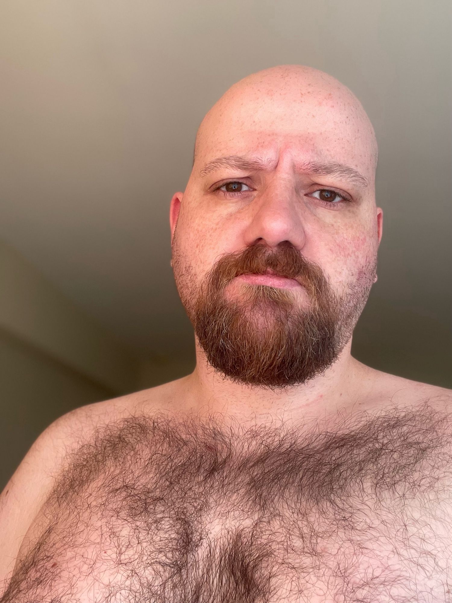 Topless man with a goatee, showcasing his hairy chest in natural lighting. He’s confidently posing with a bear pose, looking comfortable in his skin