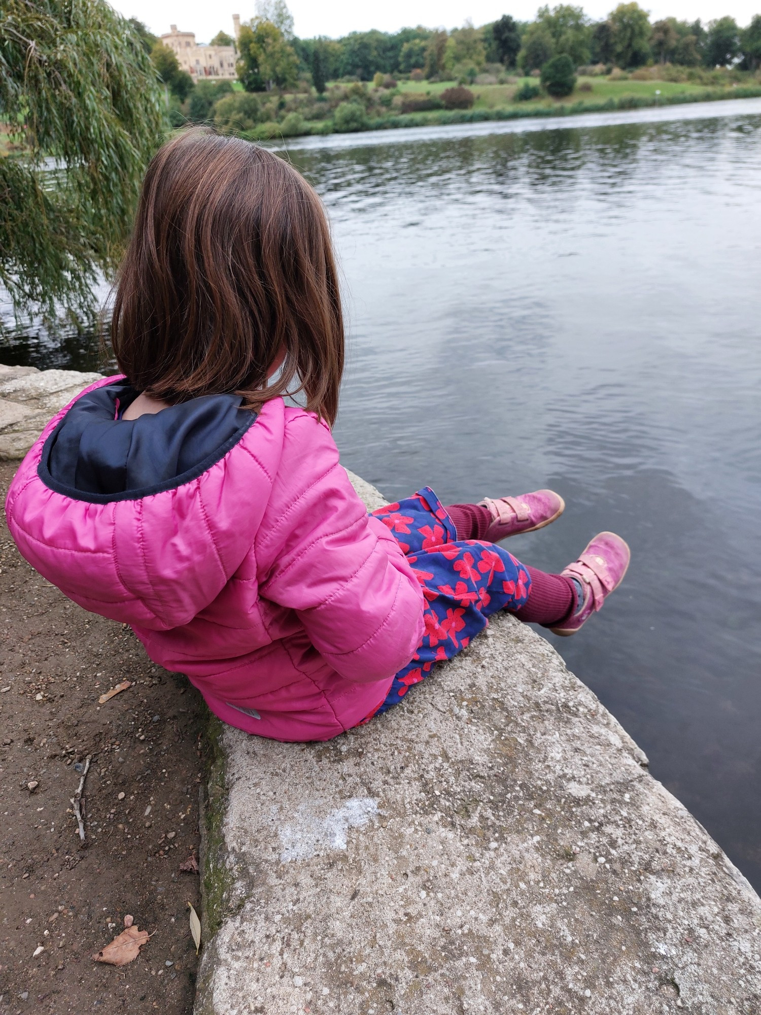 Mädchen in pinker Jacke blau roten Kleidbjnd rosa Schuhe sitzt an einer Mauer am See ubd lässt die Beine darüber baumeln