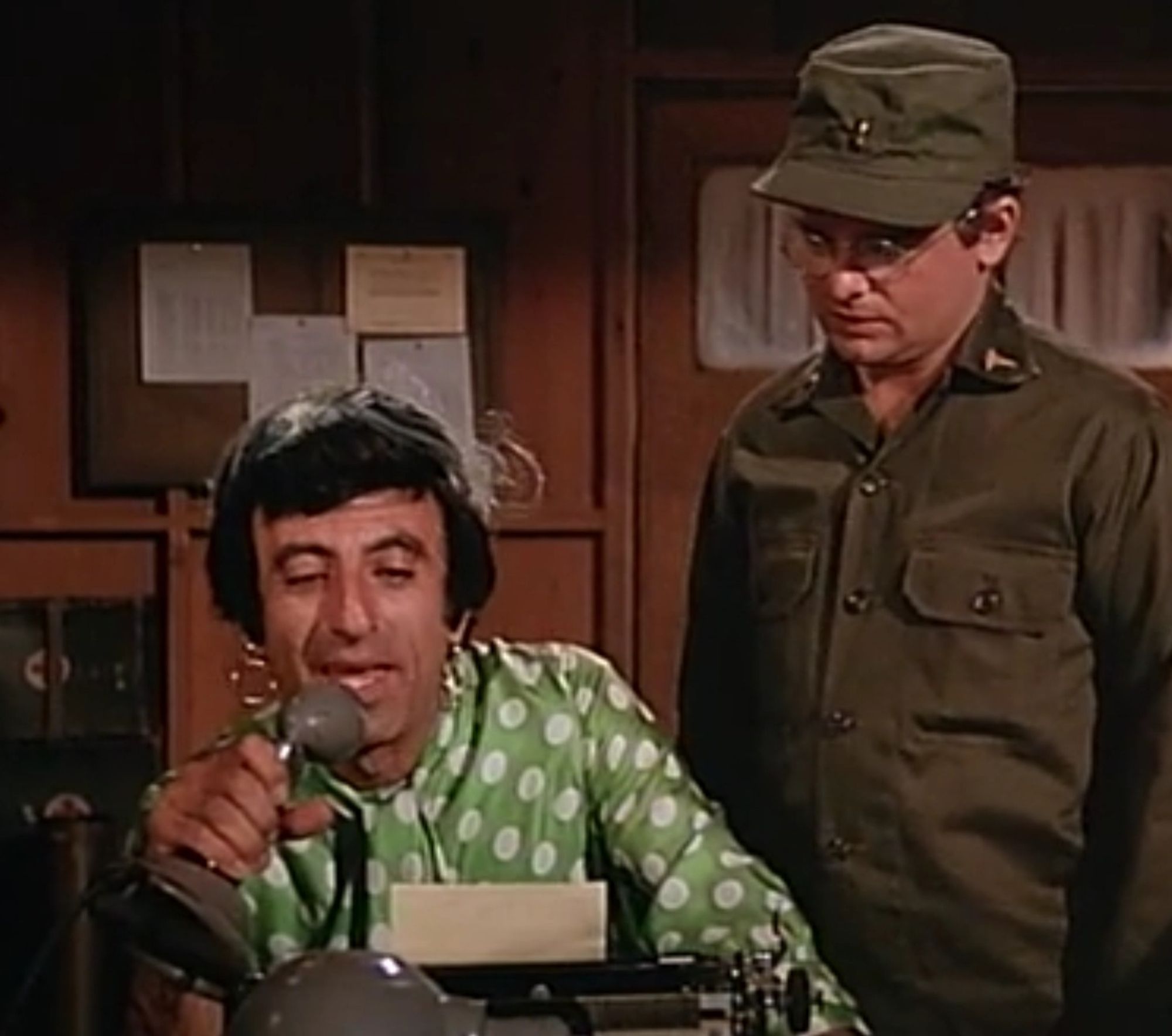 Klinger, wearing a green polka dot dress, earrings, and a darling hat, makes an announcement to the 4077 camp over the loudspeakers while Radar looks on