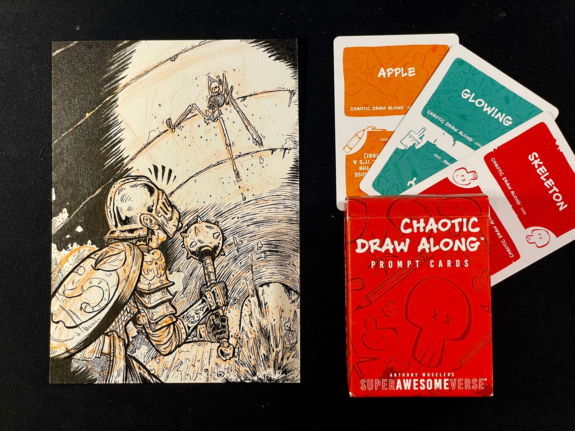 A photo of the previous illustration next to three cards and a deck box. Each card has a prompt word: "apple", "glowing", and "skeleton", respectively. The deck box reads, "Chaotic Draw Along: Prompt Cards".