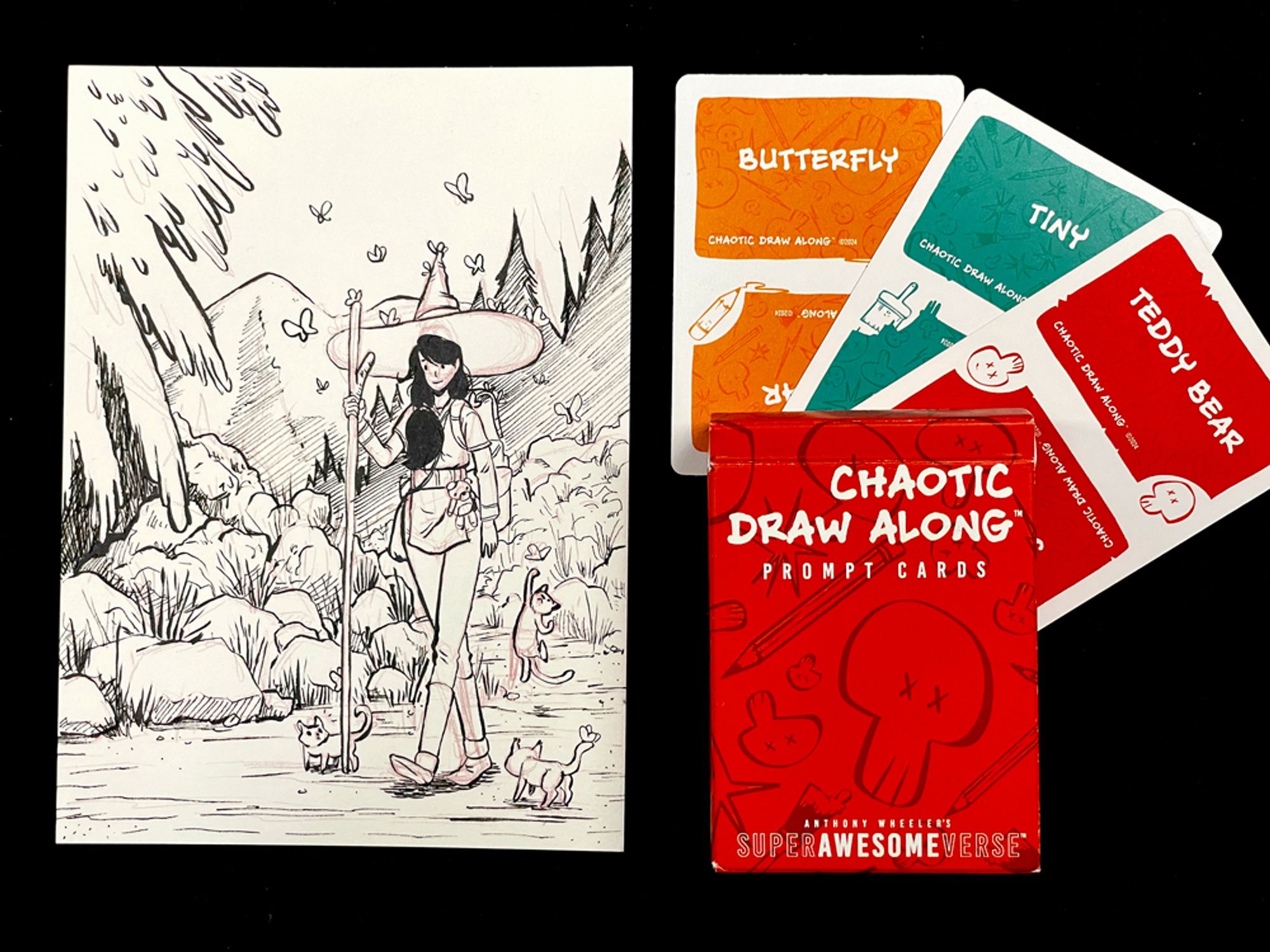 A photo of the previous illustration next to three cards and a deck box. Each card has a prompt word: "butterfly", "tiny", and "teddy bear", respectively. The deck box reads, "Chaotic Draw Along: Prompt Cards".