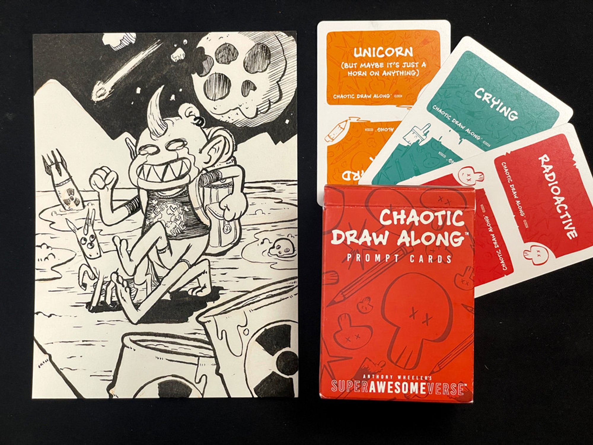 A photo of the previous illustration next to three cards and a deck box. Each card has a prompt word: "unicorn", "crying", and "radioactive", respectively. The deck box reads, "Chaotic Draw Along: Prompt Cards".