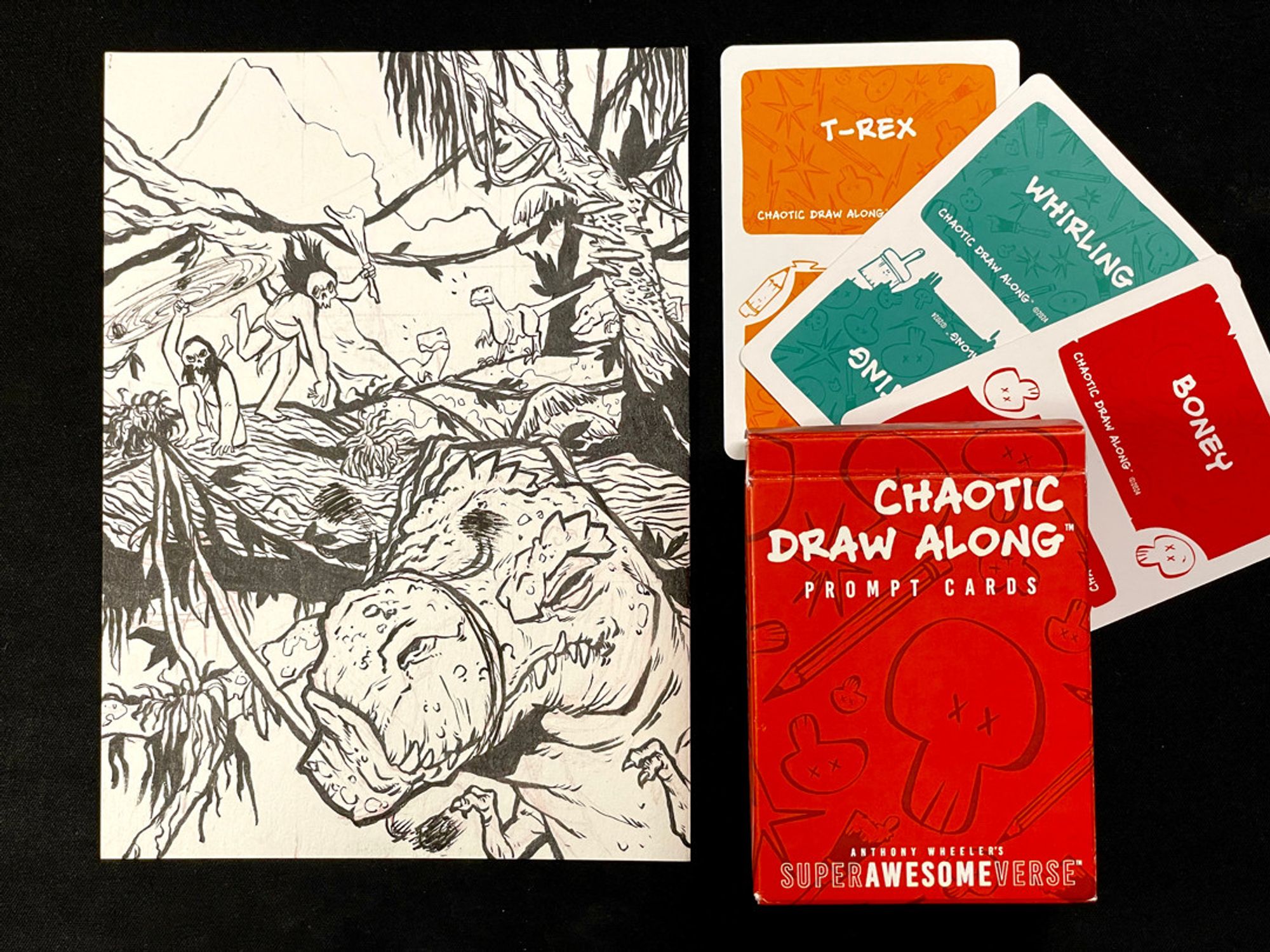 A photo of the previous illustration next to three cards and a deck box. Each card has a prompt word: "T-Rex", "Whirling", and "Boney", respectively. The deck box reads, "Chaotic Draw Along: Prompt Cards".