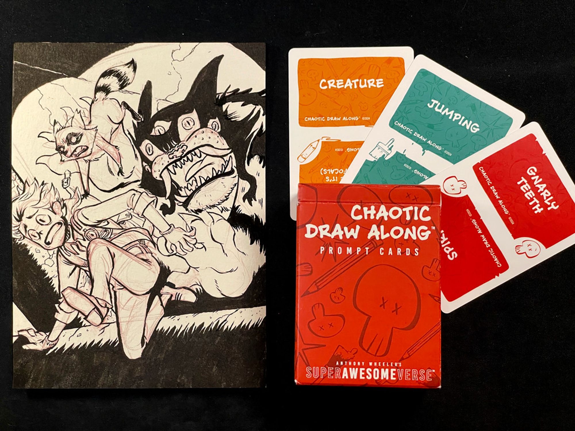 A photo of the previous illustration next to three cards and a deck box. Each card has a prompt word: "creature", "jumping", and "gnarly teeth", respectively. The deck box reads, "Chaotic Draw Along: Prompt Cards".