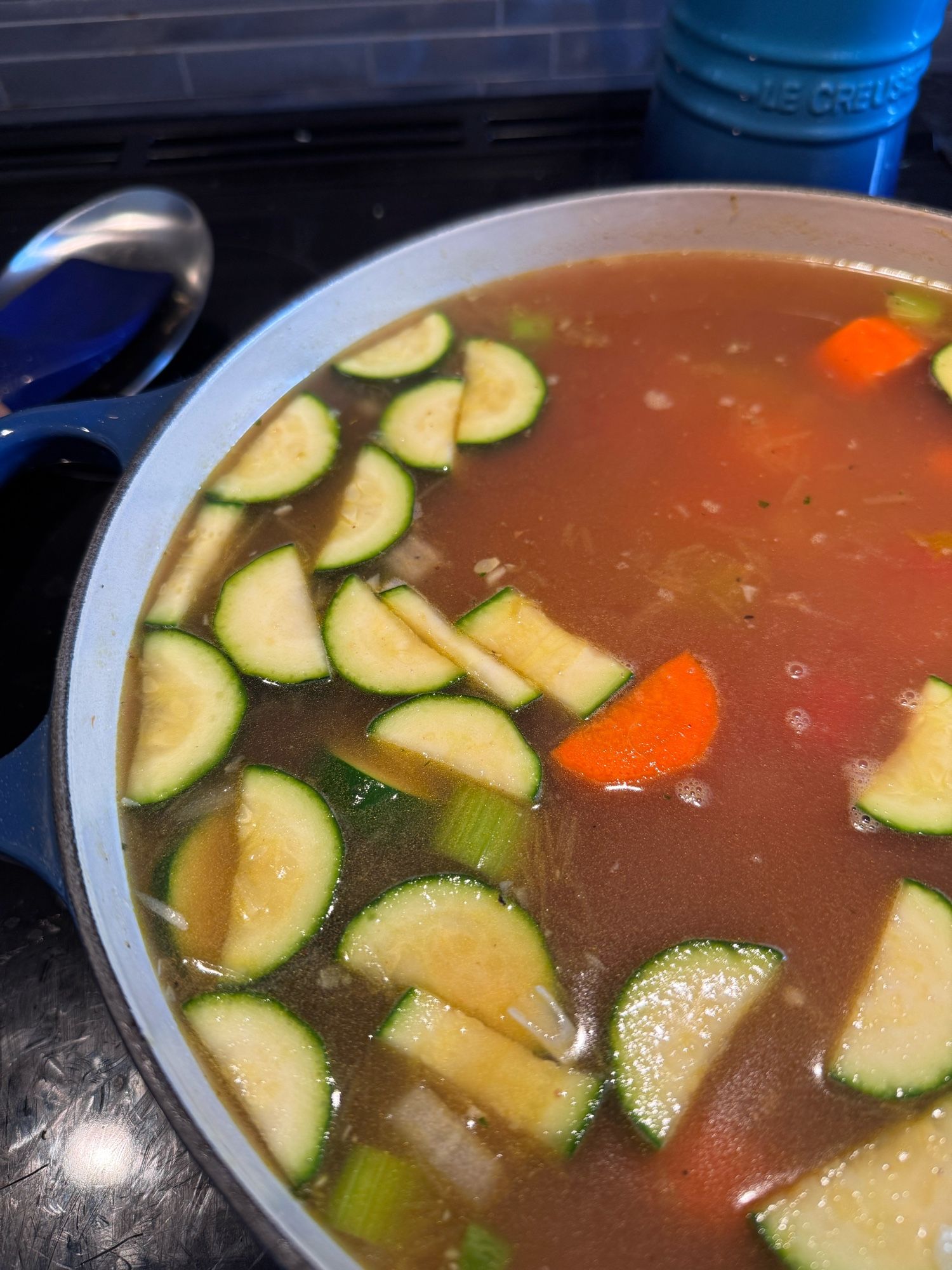 A large pot of soup simmering on a stove. Visible are the zucchini, celery, and carrot floating in a broth of chicken and tomato.