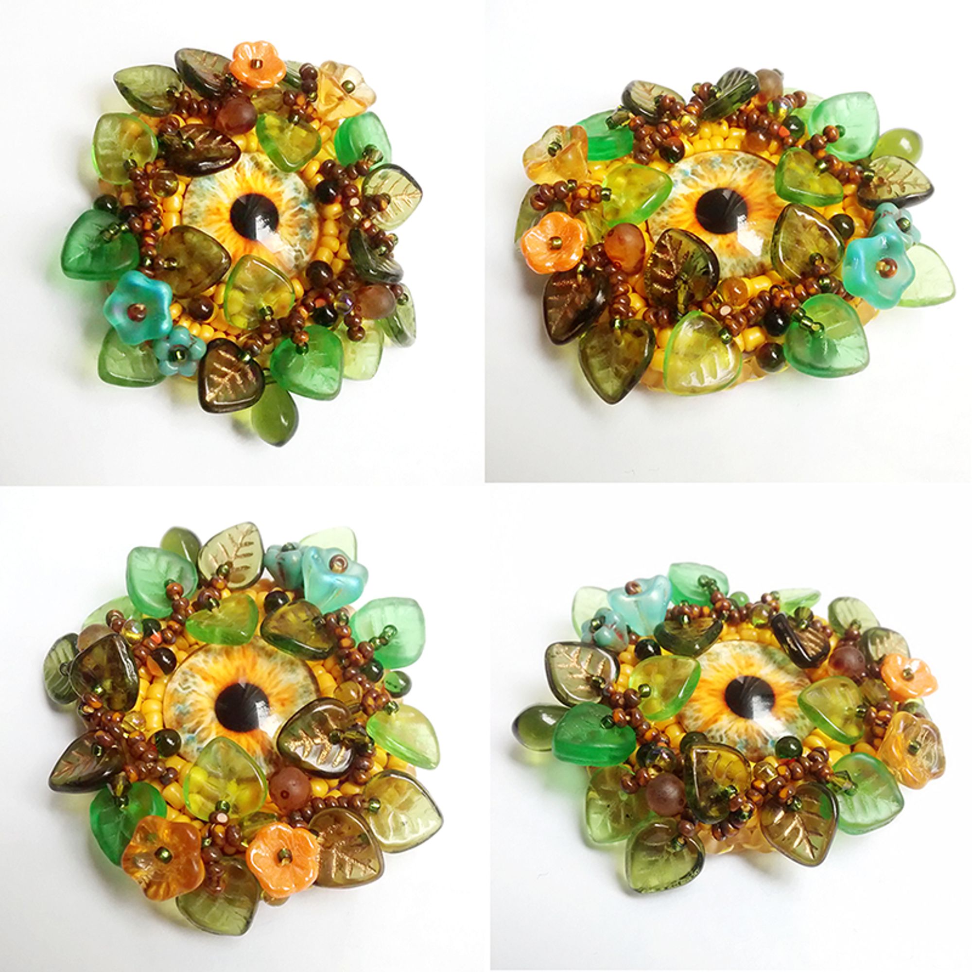 picture of a bead embroidered pendant or brooch, featuring an eye in yellow shades hiding behind a flurry of translucent glass leaves, berries and flowers in green and autumnal shades.