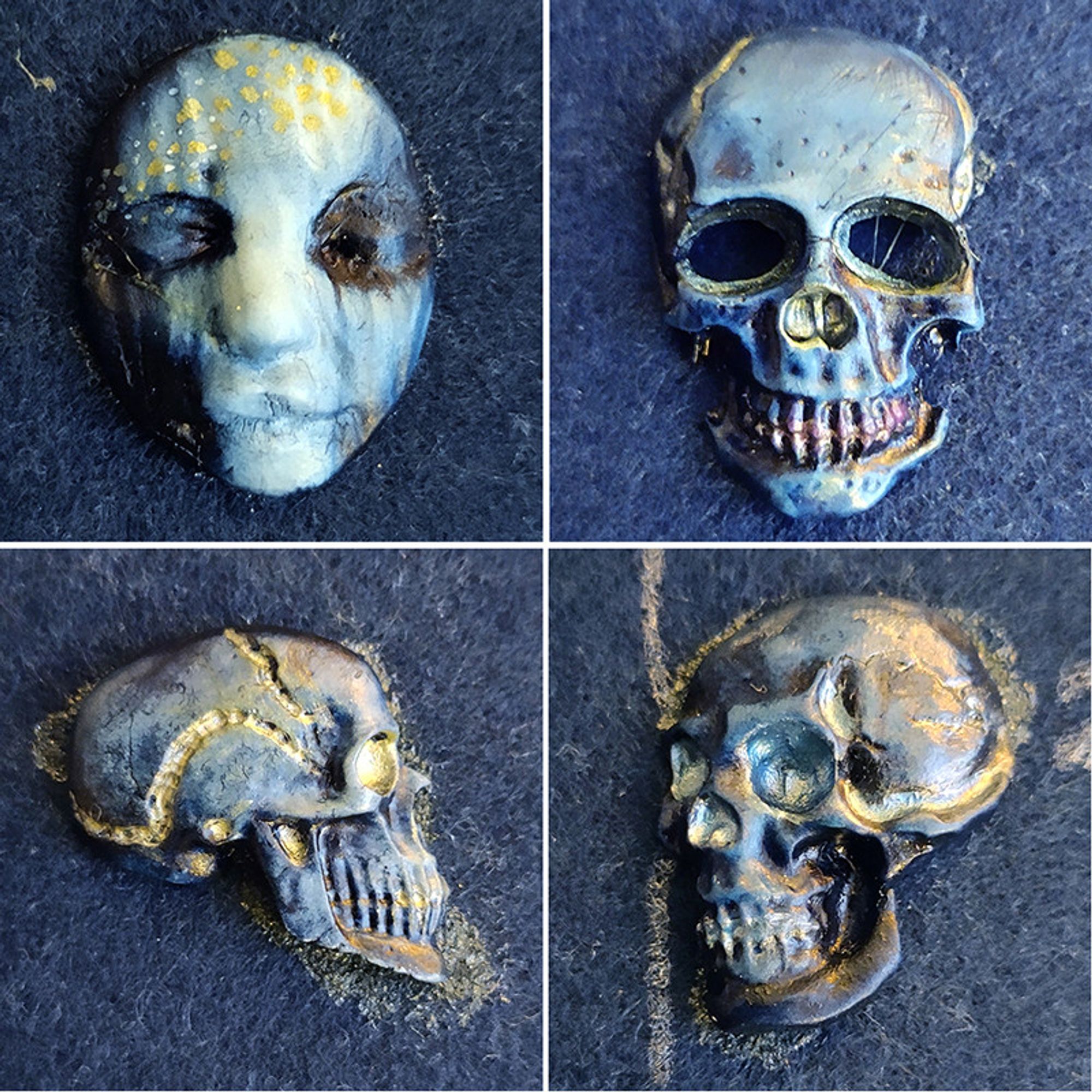 close up of a fimo pale face cabochon with closed eyes and 3 skull fimo cabochons, in shades of blue and black with golden or silver blue accents on the forehead, glued to a dark blue felt surface.