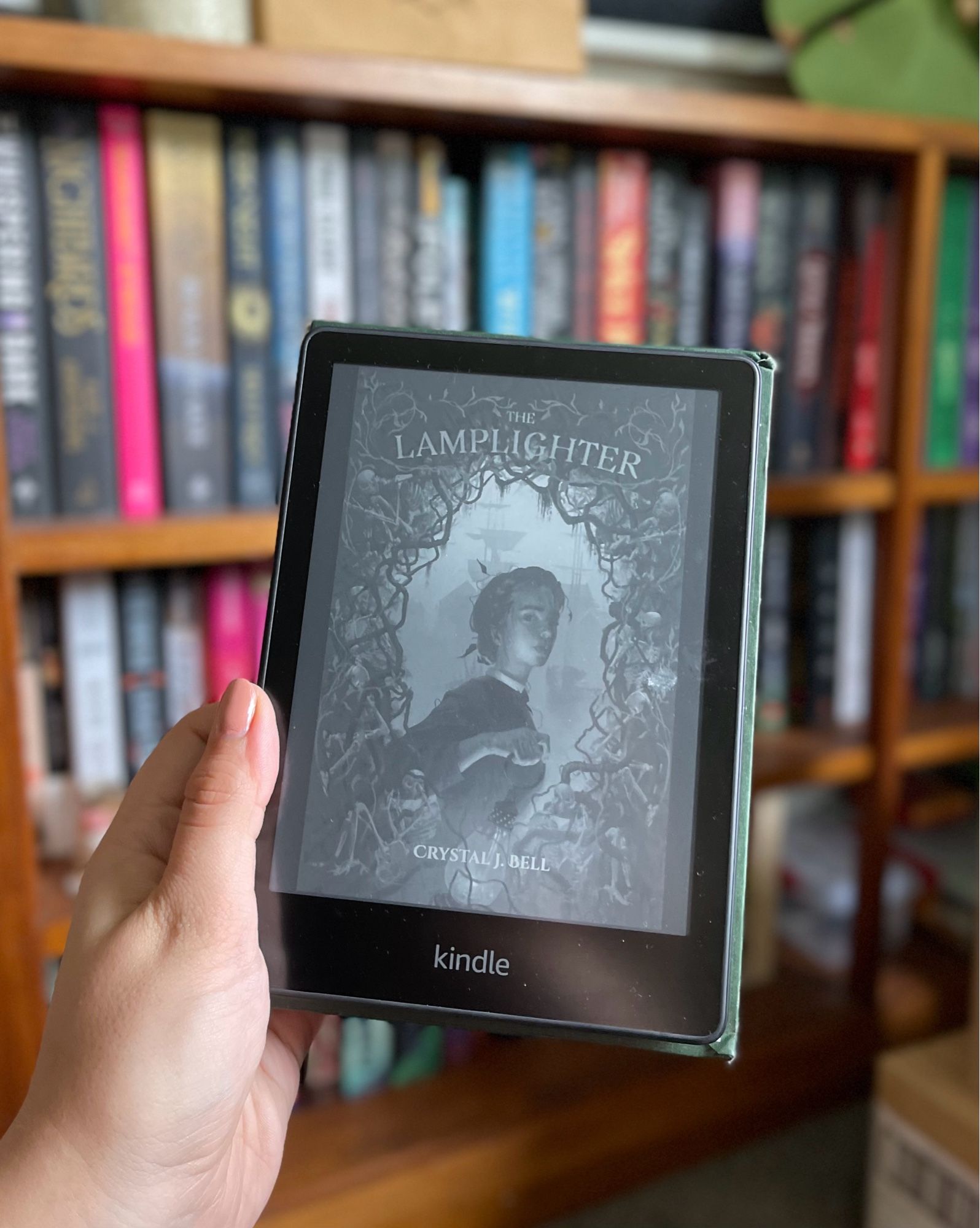 I’m holding my kindle with the cover of Lamplighter by Crystal J. Bell on display. There is a colourful bookshelf in the background, slightly blurred.