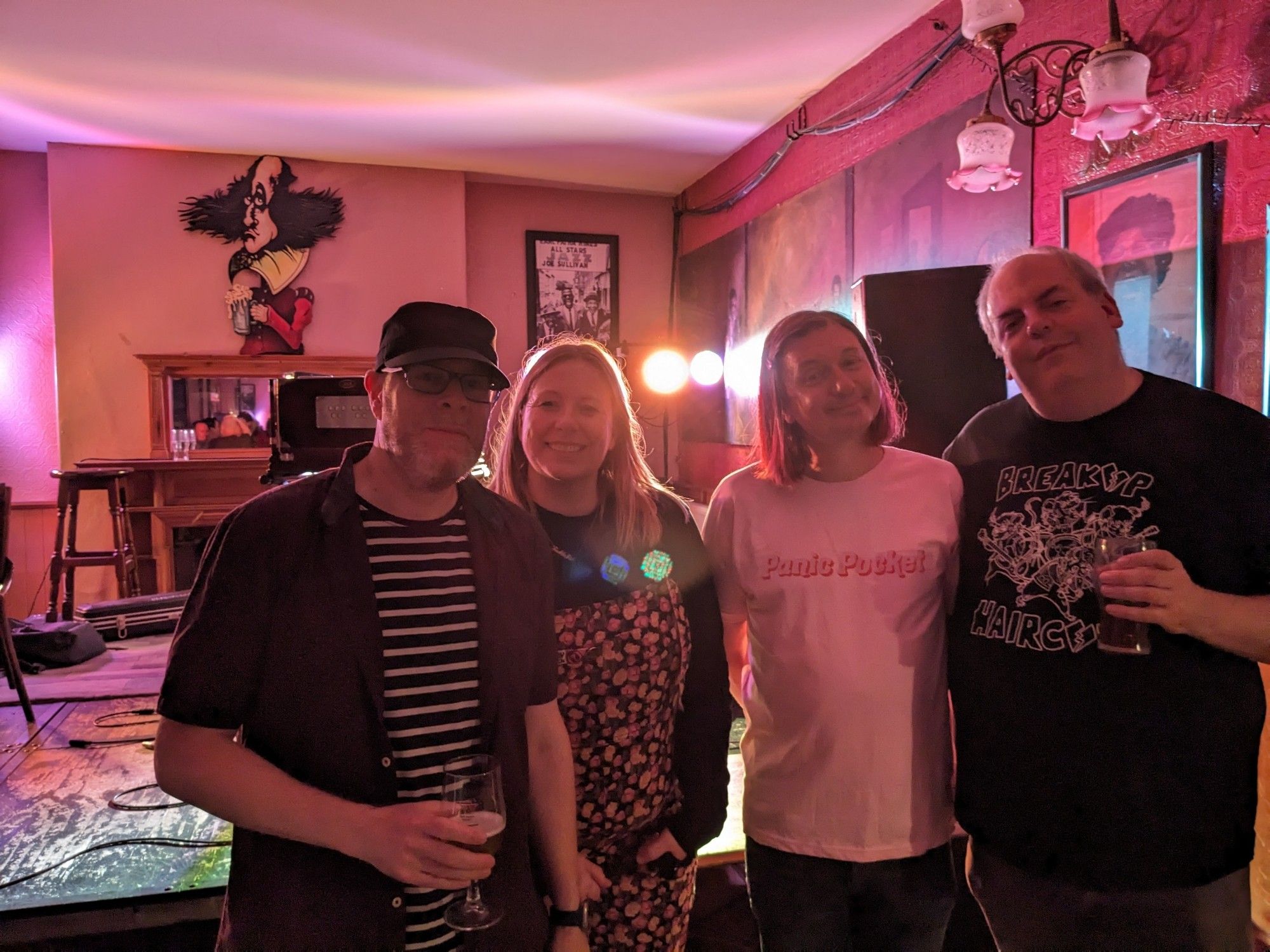 Four tired but happy people smiling into the camera with the upstairs room at Shakespeare's in the background - the stage and pictures on the wall