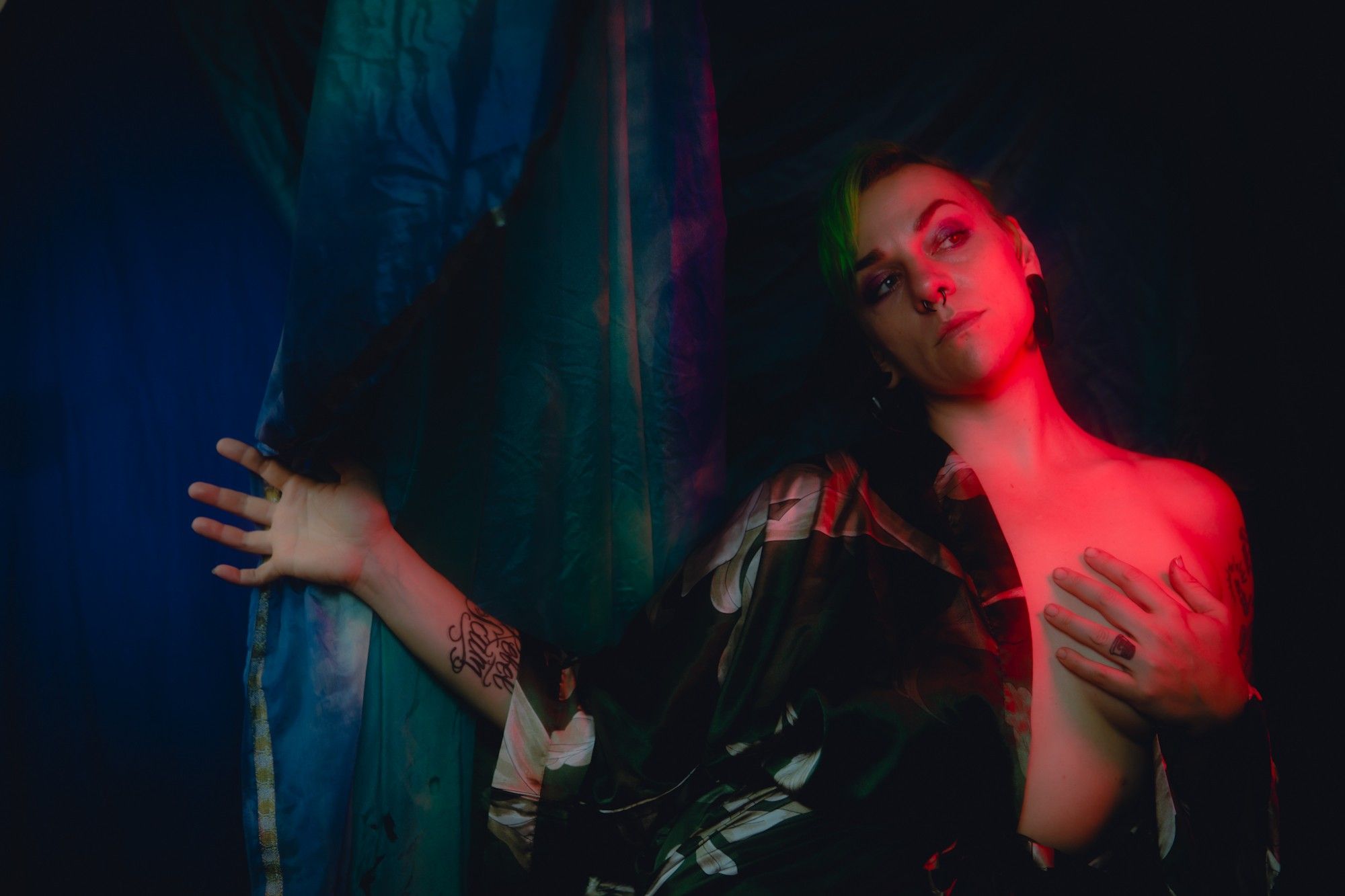 A photo of Gemma Raelit limned in vivid red tones, 
A brow arched as she faces the camera but her eyes are cast to the side, a silk robe falling off her left shoulder with her left hand covering her breast and her right hand outstretcched to hold back a curtain of fabric. The background is drapes of teal and ultramarine fabric, fading unto the darkness.