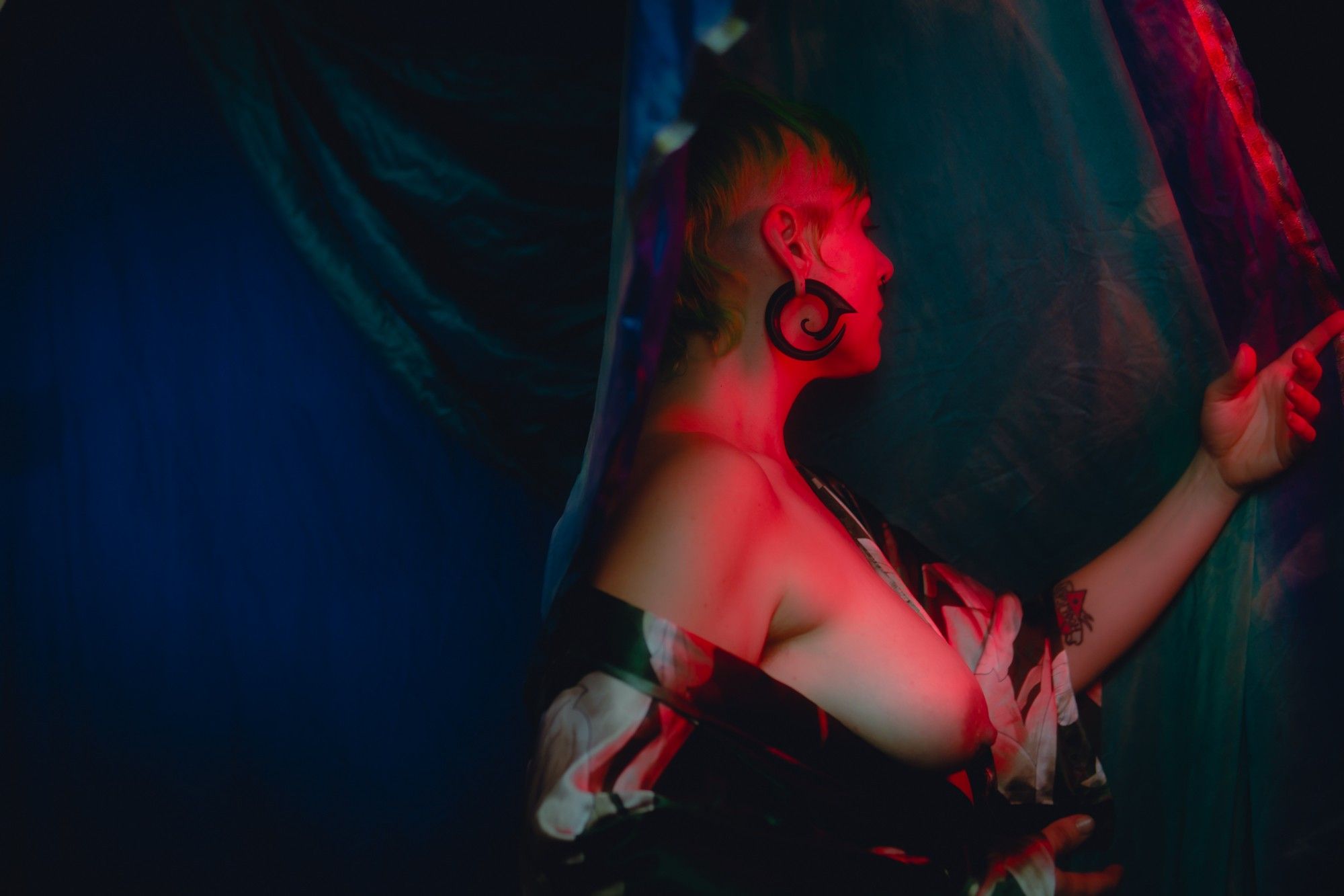 A photo of Gemma Raelit limned in vivid red tones, 
she is just turned away from the camera with her profile only just visible, a silk robe falling off her left shoulder bares her breast and her right hand is outstretched to hold back a curtain of fabric. The background is drapes of teal and ultramarine fabric, fading unto the darkness.