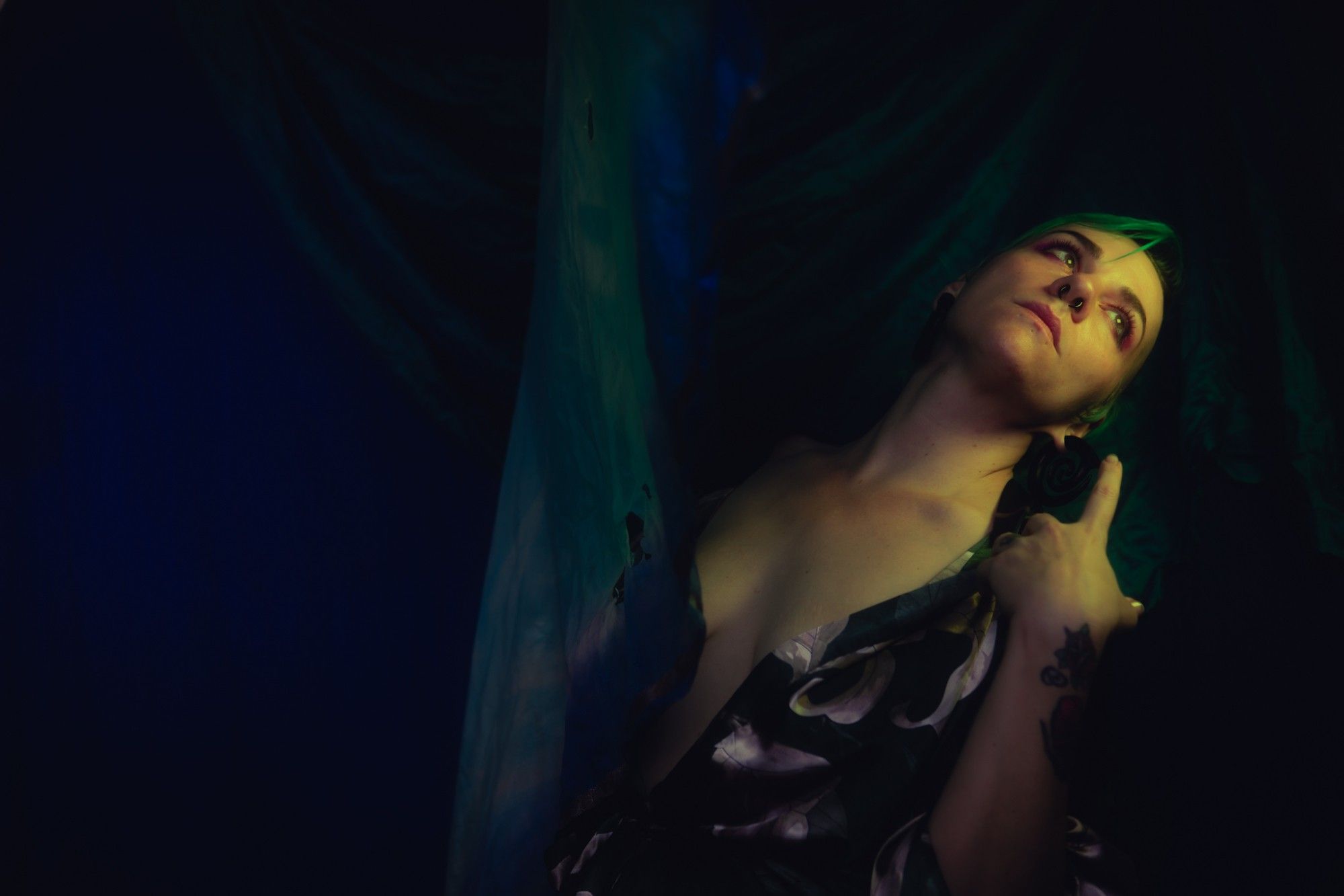 A photo of Gemma Rae lit in soft green-gold tones, she's leaning back, her head tilted up and to the side as she gazes towards the ceiling, a silk robe open at her sternum. The background is drapes of teal and ultramarine fabric, fading unto the darkness.