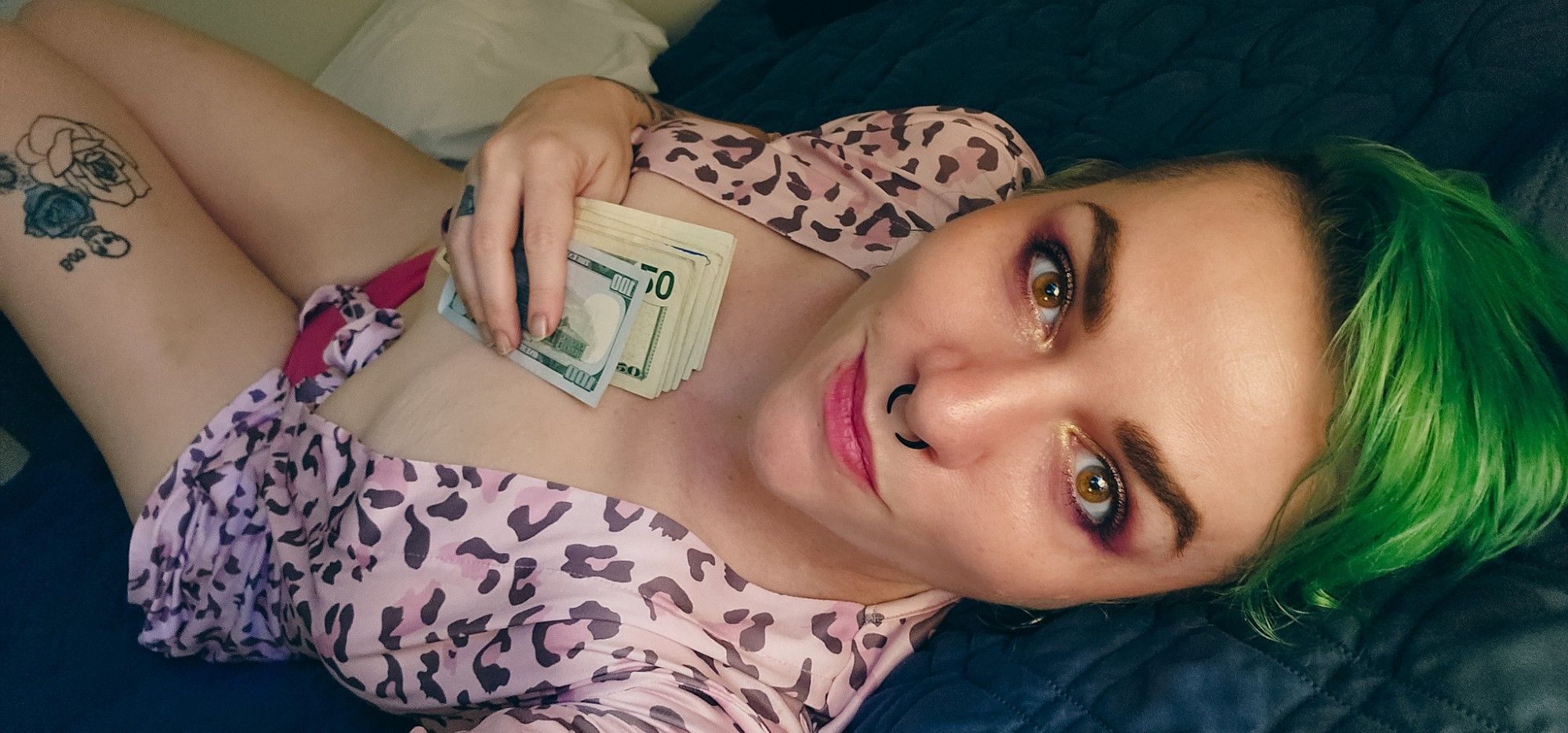 A selfie of Gemma Rae, a femme with green hair and a septum ring, laying down on a dark blue blanket, wearing a pale pink leopard print robe that's open at the sternum and bright pink panties. They're looking up at the camera with a slight smile and clutching a stack of bills to their chest.