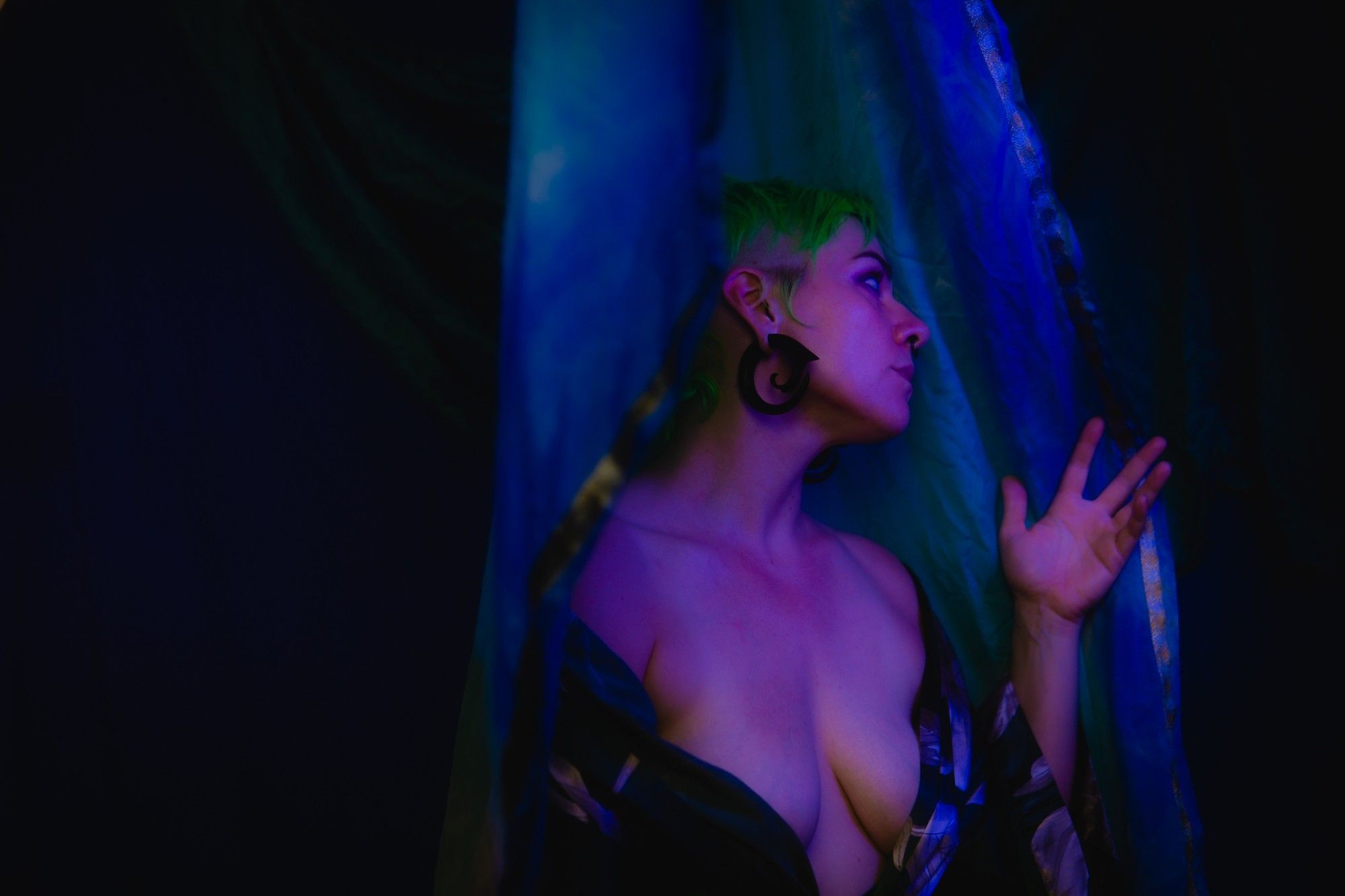 A photo of Gemma Raelit in soft magenta tones, she's in side profile, staring into the distance, a silk robe falling off her shoulders and just covering her nipples. The background is drapes of teal and ultramarine fabric, fading unto the darkness.