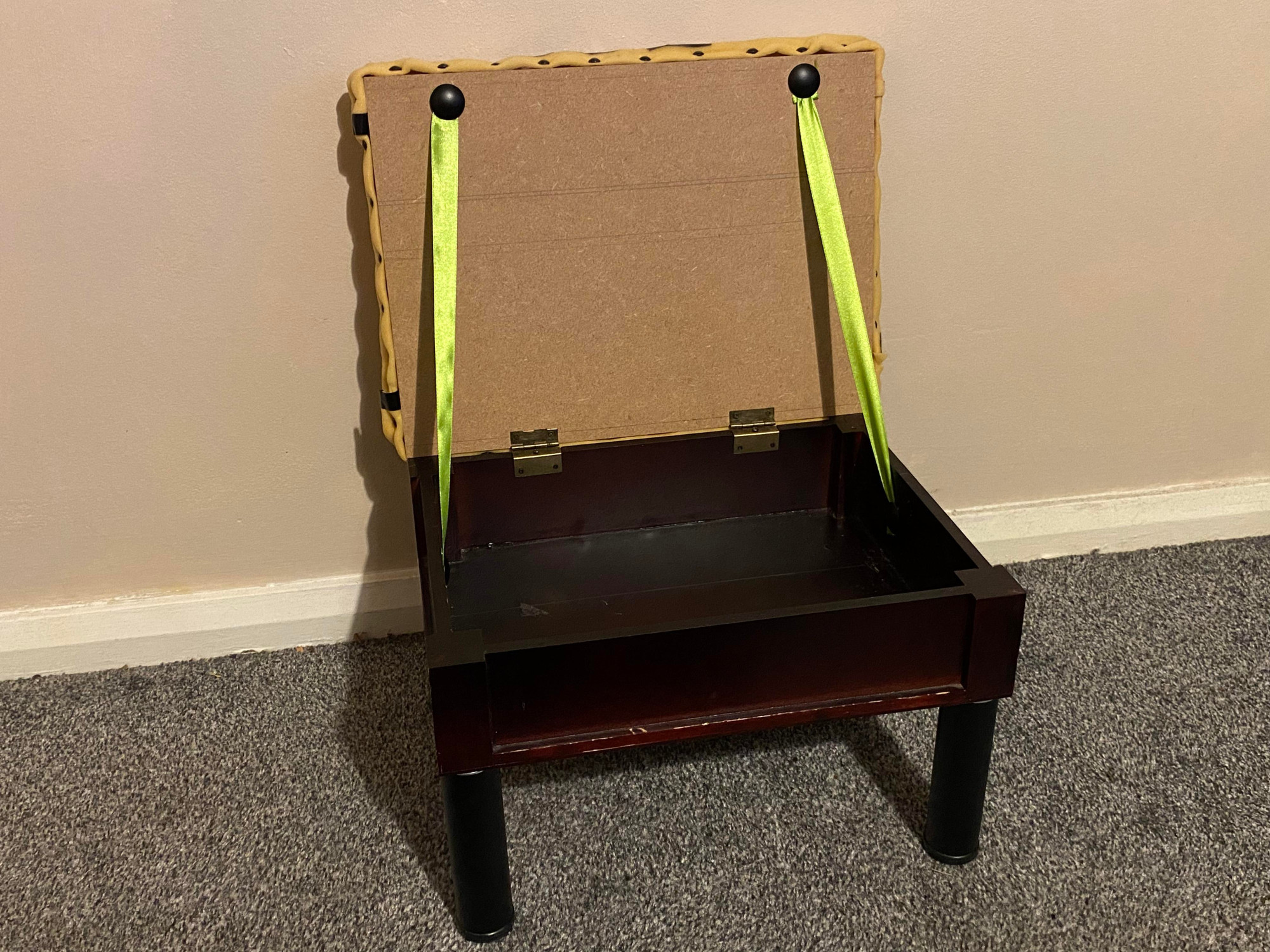 Footstool with the storage lid open. There are two lid supports made from green ribbons now attached to it. Front view.