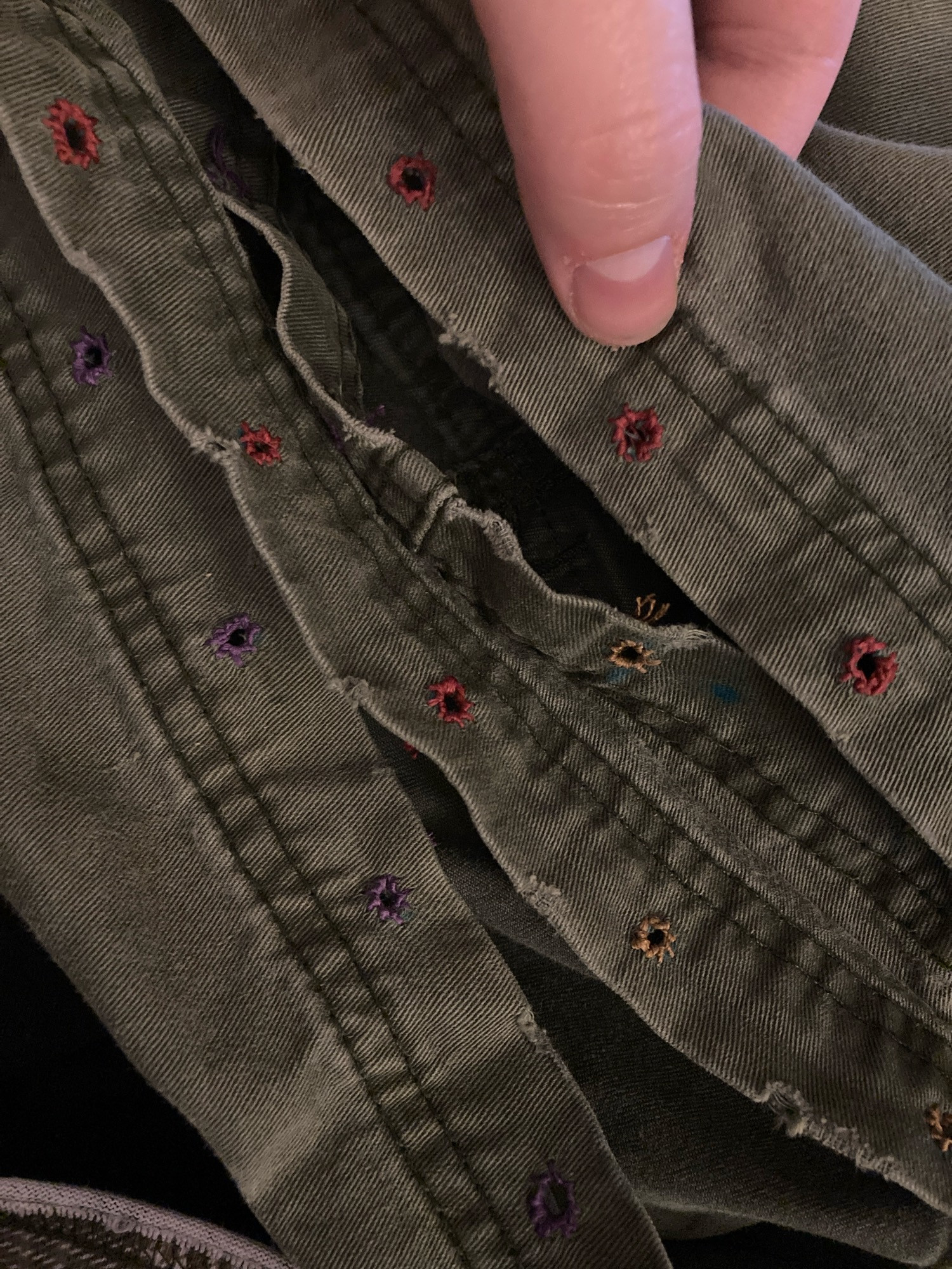 an up-close photo of the bottom hems of a pair of olive green shorts. Cody’s thumb holds down the top of one edge (for scale), showing rows of multicolored embroidery thread sewn into holes, making tiny eyelets. 
