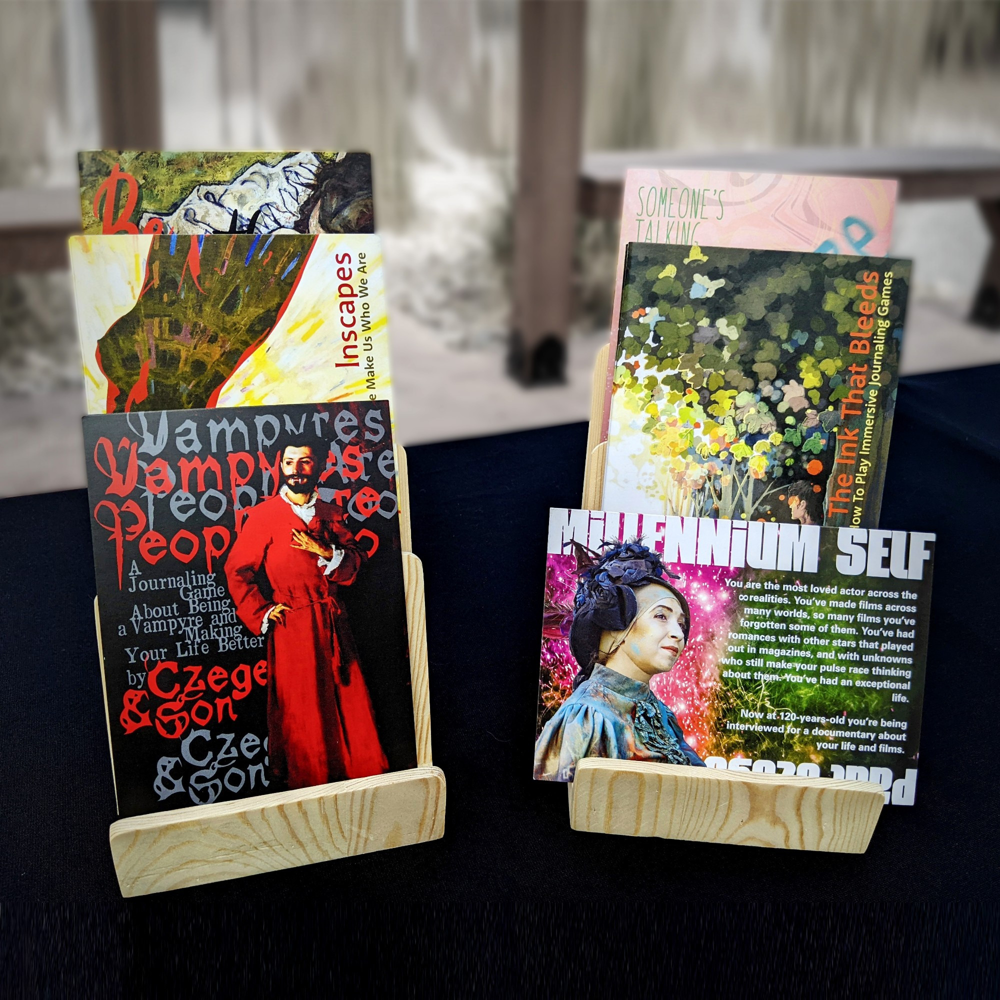 Two wooden zine displays showing copies of The Ink That Bleeds, Inscapes, Be With Me, The Sneeze, Vampyres Are People Too, and Millennium Self .
