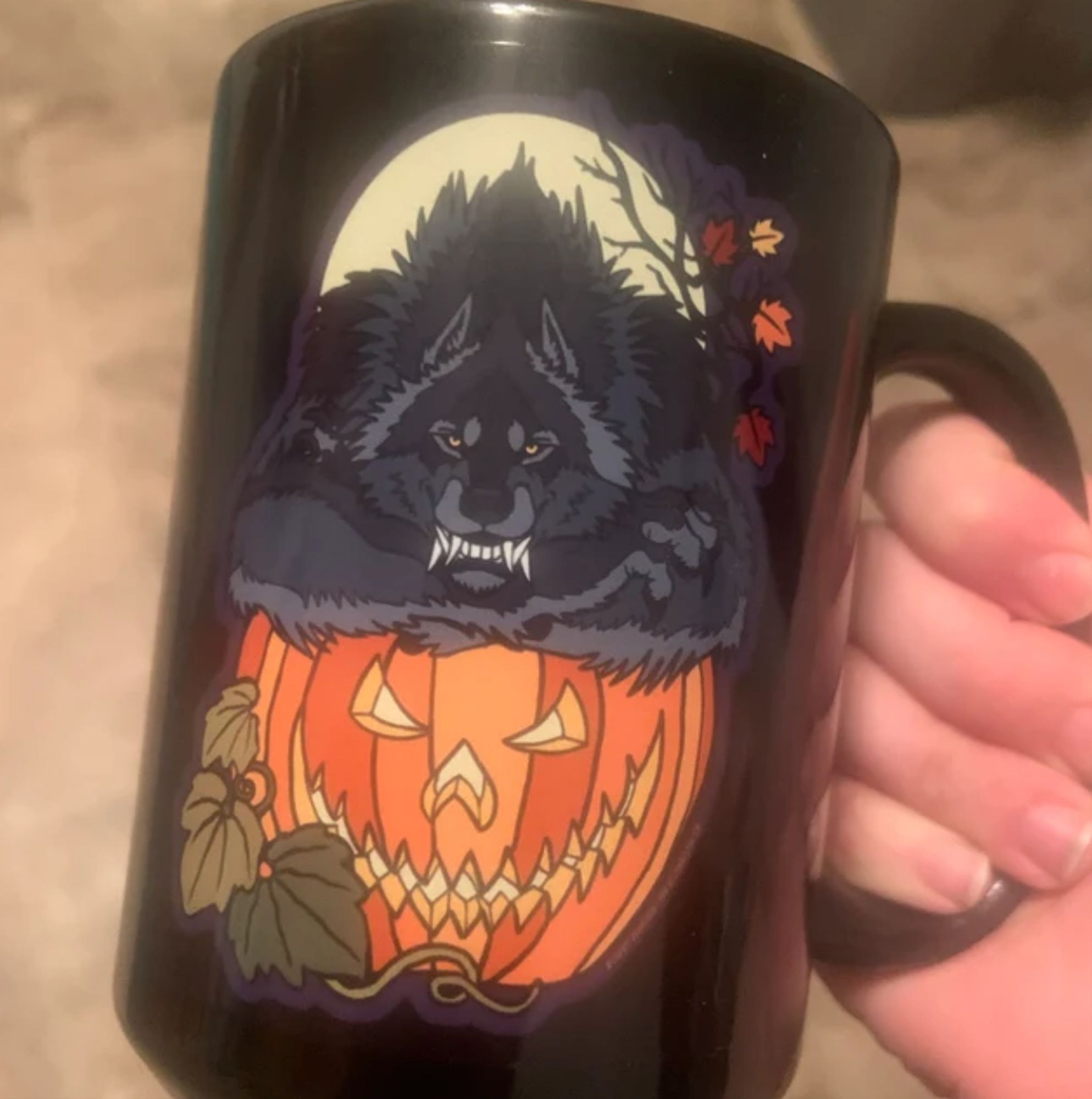 Photo of a hand holding a large black coffee mug with a Jack o'lantern and a grinning werewolf printed on it