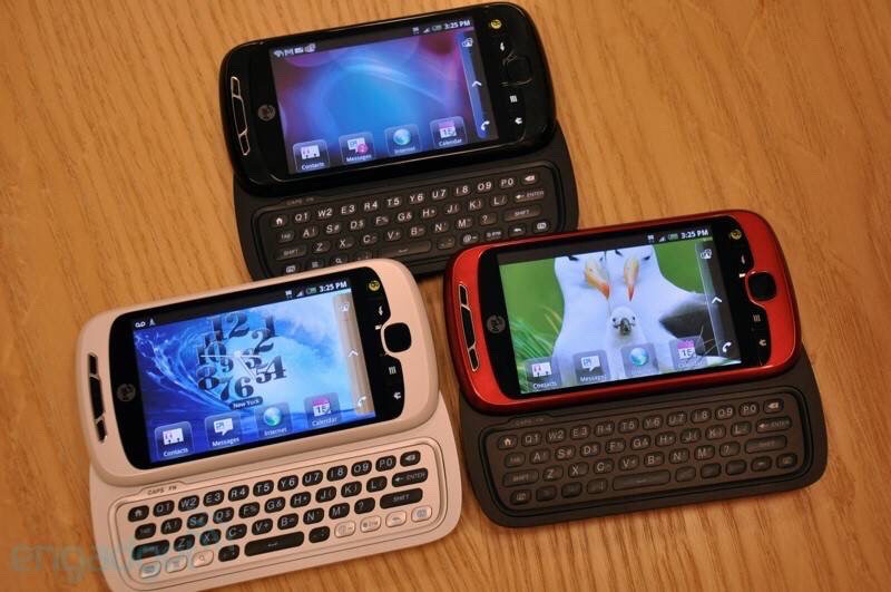 A wooden table has three T-Mobile HTC My Touch 3G slide out querty keyboard touchscreen cellphones laughing open in it. A black, a white, and a red. The red phone has a seagull family as the screen background.