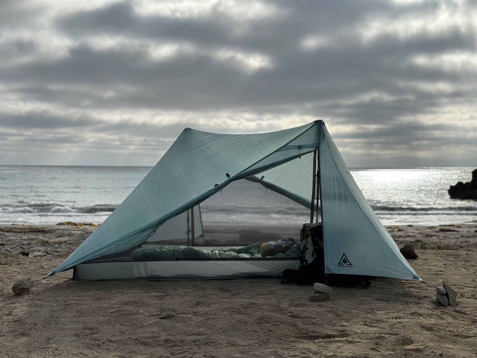 A tent is set up on the beach in front of the ocean. The doors are open and you can see the waves through the tent. A moody sky is overhead.