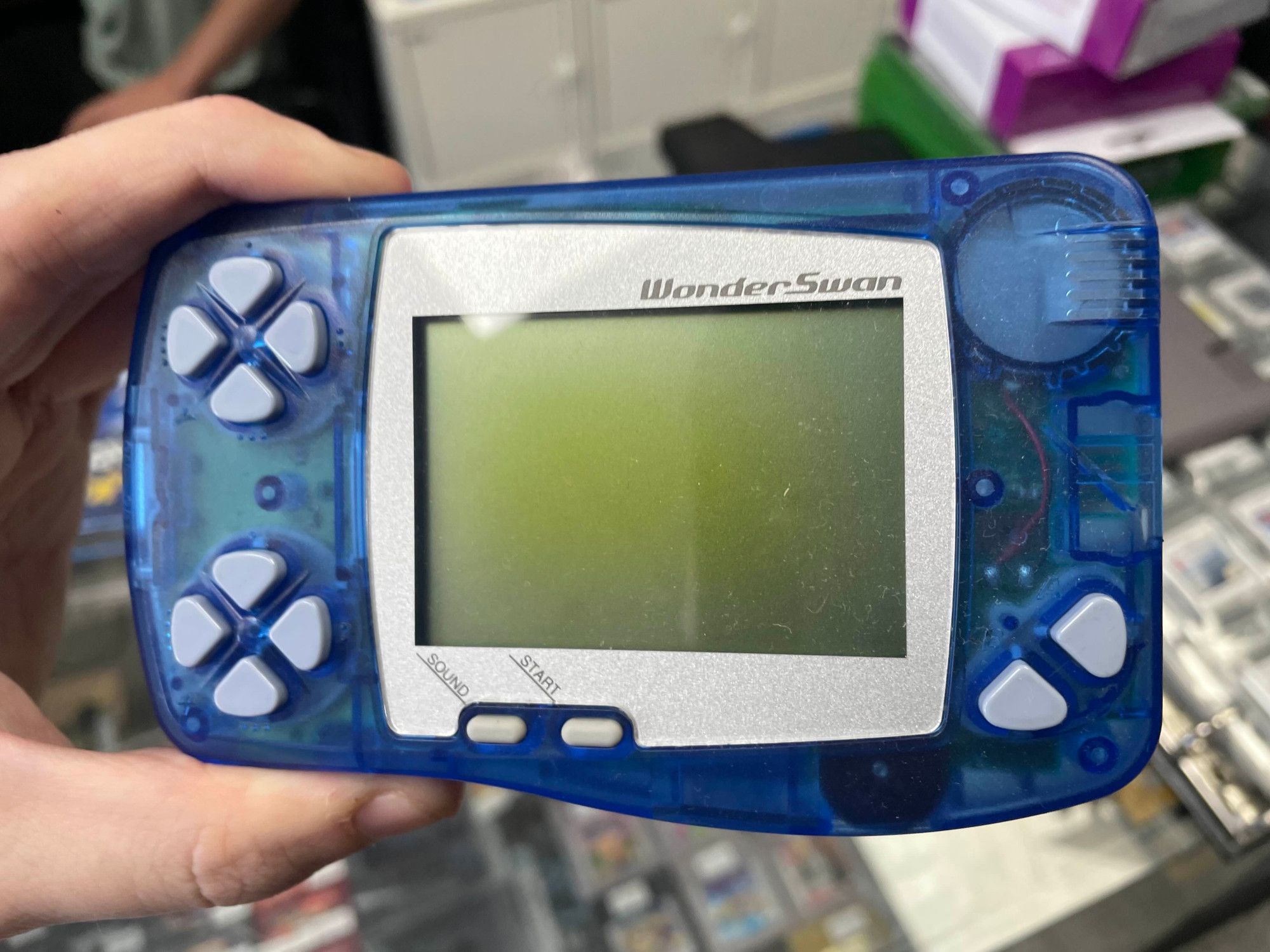 Me holding a Bandai WonderSwan, a handheld game console released in 1999. It has a monochrome screen, A and B buttons, as well as two whole sets of four buttons labelled X and Y. There's also "sound" and "start" buttons. The transparent blue shell allows you to see some of the inner workings of the console.