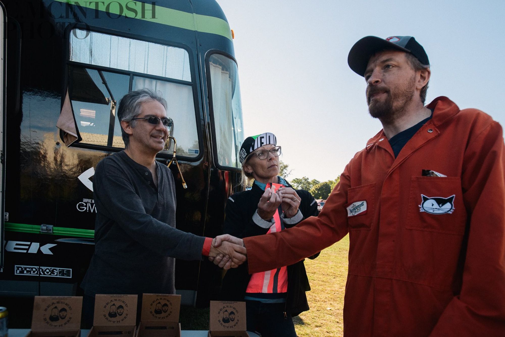 Three people in front of the Durrin Team Motorhome, two men shaking hands, each looking in a different direction and a woman in a cycling cap with a mouthfull of food looking in a third direction.