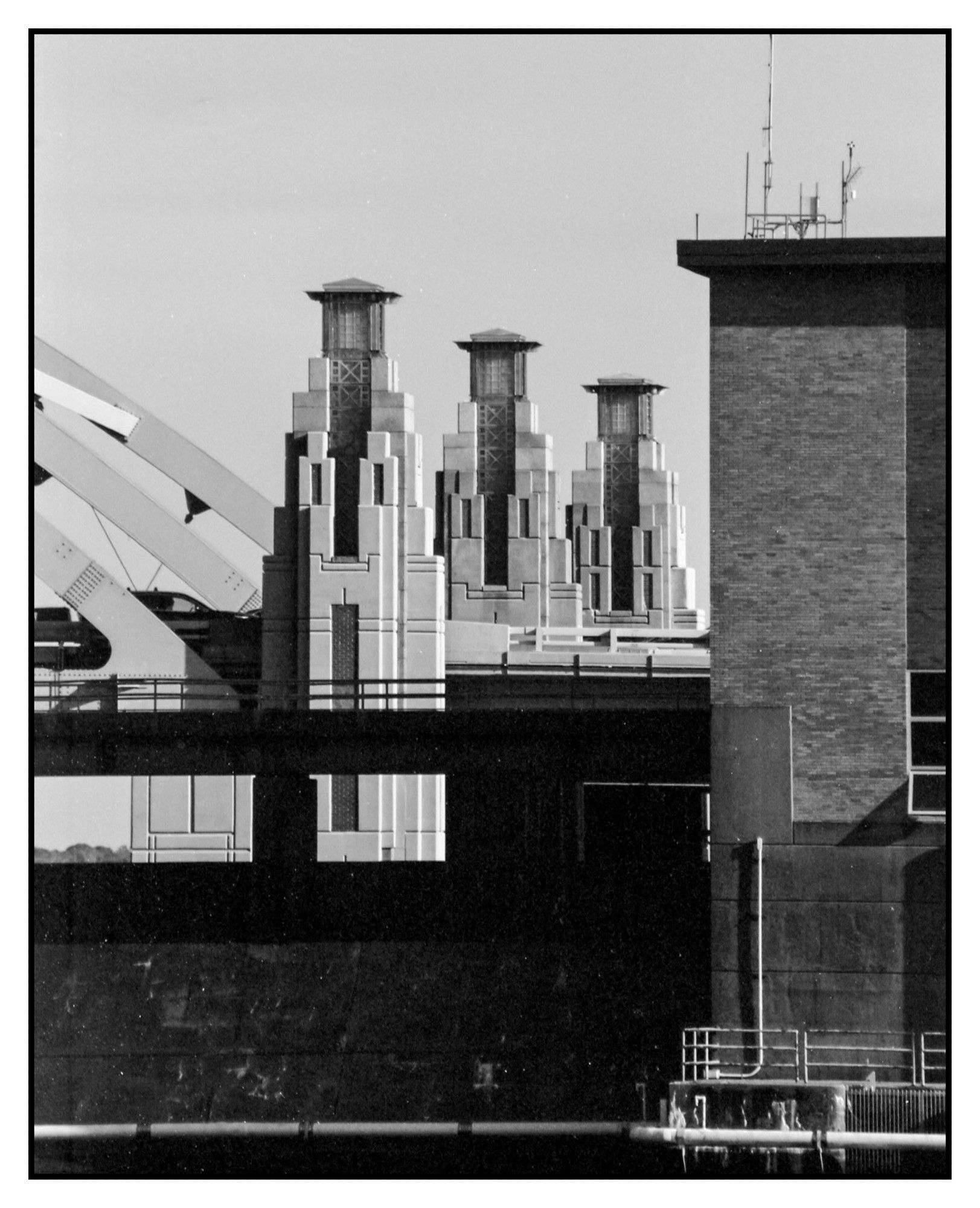 three towers on the bridge with a brick building on the right and the beginning of the arches on the left.