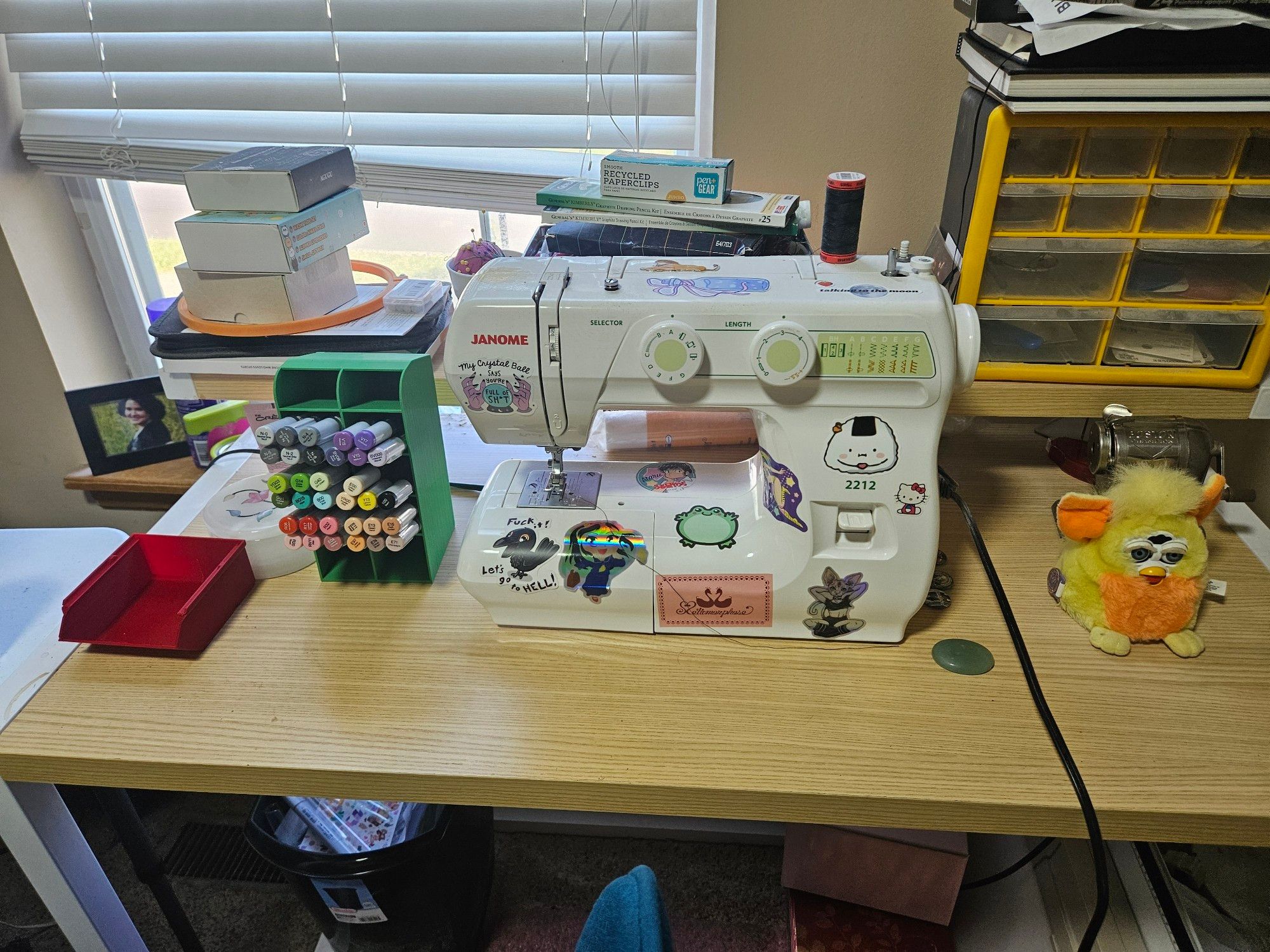 A table with a sewing machine covered in stickers, a storage shelf of copic refills, and a random baby furby.